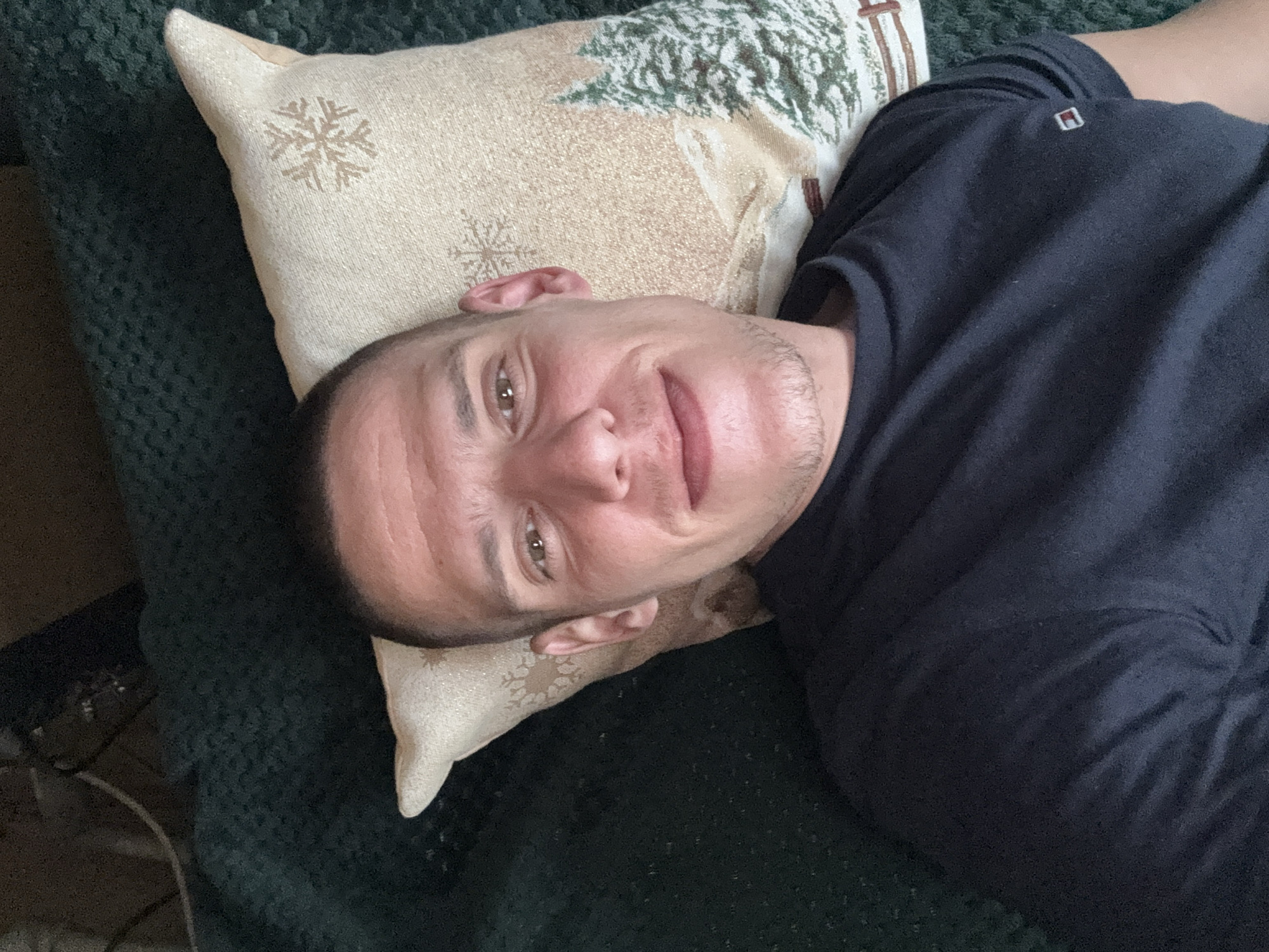 A man is lying on a dark green textured surface, resting his head on a beige pillow decorated with snowflakes and a Christmas tree design. The man is wearing a dark blue shirt with a small logo near the shoulder. The location is not discernible.