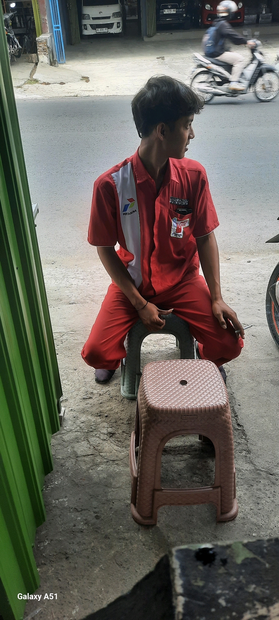 In Selopuro, Indonesia, a young male employee wearing a red uniform with white accents and the "Pertamina" logo sits on a light gray plastic stool. He is holding a cigarette in his right hand. In the background, a white van with the license plate AG 8781 HA is visible, along with a black car with license plate N 1370 RH. A motorcycle with a rider wearing a helmet is in motion on the road. A green corrugated metal structure is on the left side of the frame. Another brown plastic stool is placed in front of the stool the man is sitting on. The ground is made of concrete and asphalt. The overall setting appears to be a street or roadside area in a commercial district.