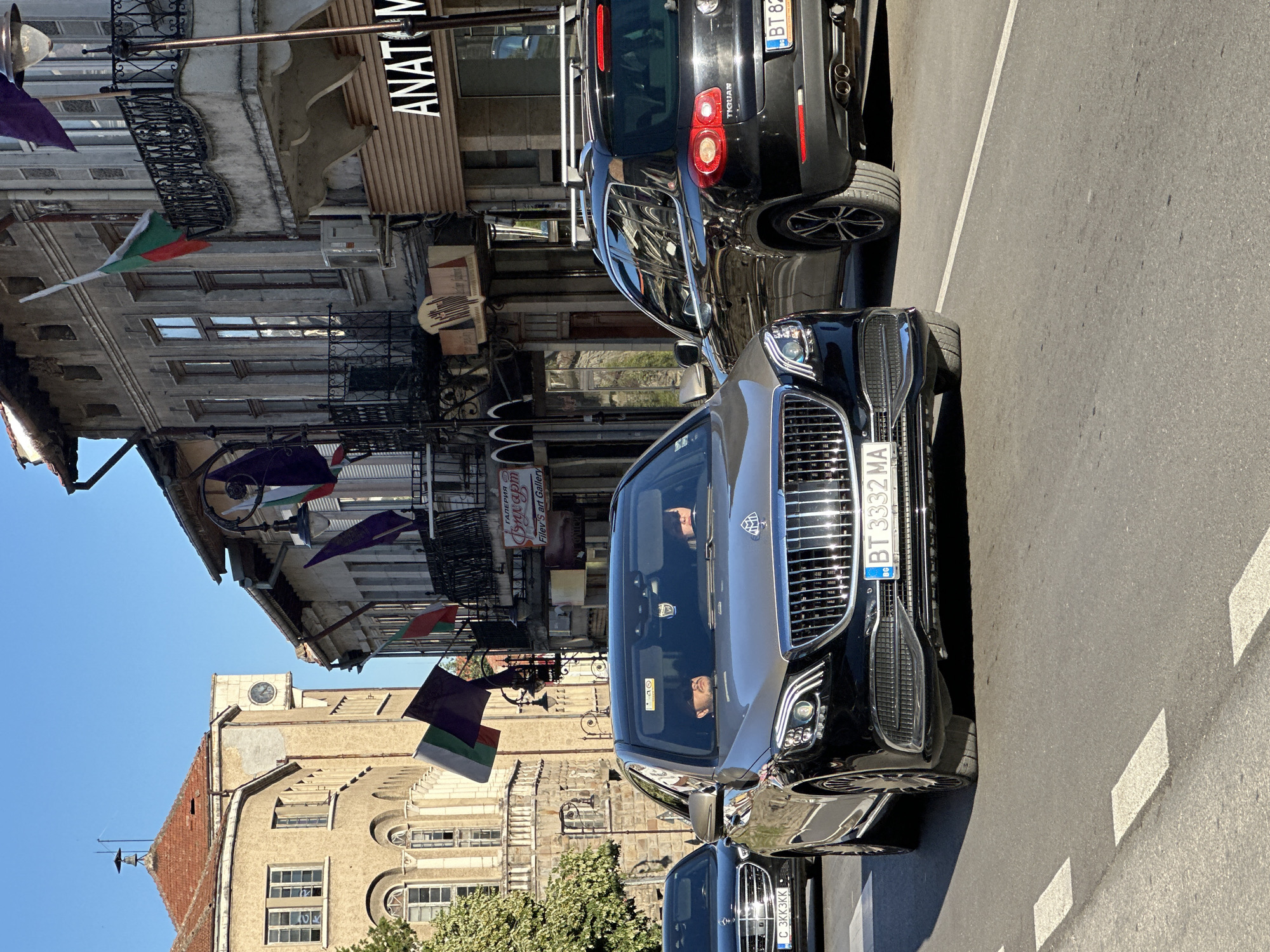 The image captures a street scene in Veliko Tarnovo, Bulgaria, under a clear blue sky. In the foreground, a luxurious black and silver Mercedes-Maybach sedan drives down the street, its license plate reading "BT 3332 MA". A black Volkswagen Tiguan SUV is positioned to the right of the Mercedes-Maybach, its license plate starting with "BT 82". A black Mercedes-Benz sedan with the license plate "C 3KK3KK" is visible in the left background.
The street is lined with buildings exhibiting traditional Bulgarian architecture, adorned with Bulgarian flags and purple flags. One building features a prominent clock tower. Several storefronts are visible, including one with the sign "ANATOMY". The overall impression is that of a bustling urban environment on a sunny day.