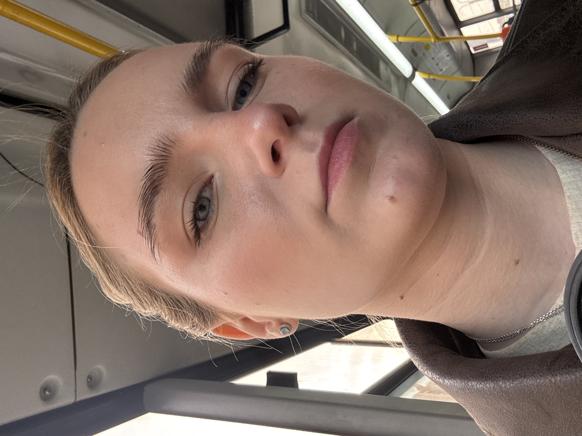 A close-up, rotated image shows a fair-skinned young woman with light blonde hair and grey-blue eyes. She has well-defined eyebrows and subtle pink lipstick. A small, circular earring is visible in her left ear. She is wearing a brown leather jacket over a light-colored shirt, and a thin silver chain necklace. The background reveals the interior of a bus, featuring a yellow handrail, overhead lighting, and a bus window. Location is Unknown, Unknown.