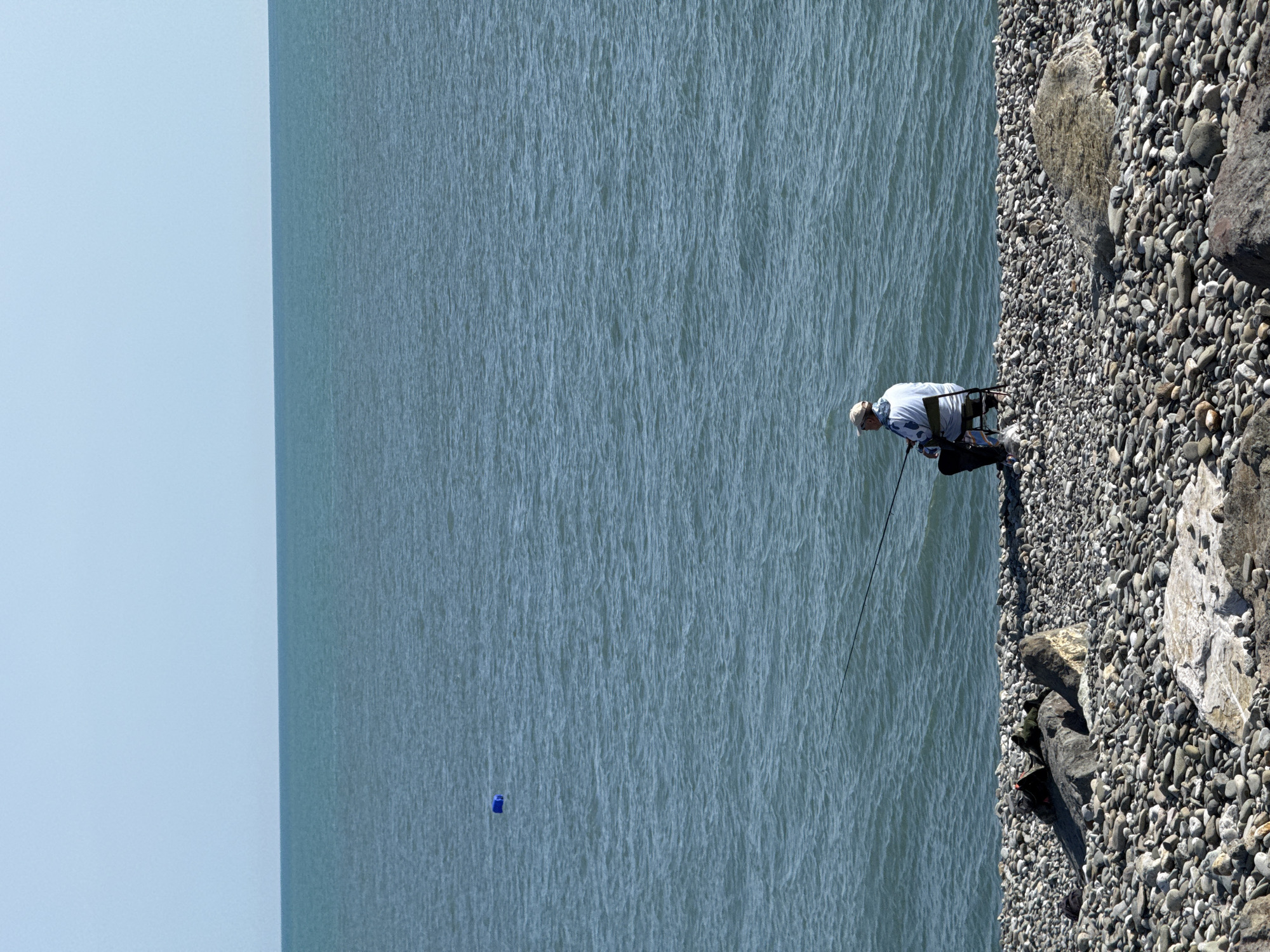 An eye-level shot, taken in the city of Sochi, Russia, shows a person fishing in the sea. The person is standing on a rocky shore, and is wearing a light-colored shirt and pants, and a baseball cap. The fishing rod extends out over the water. A blue object floats in the water. The sky is clear and blue.