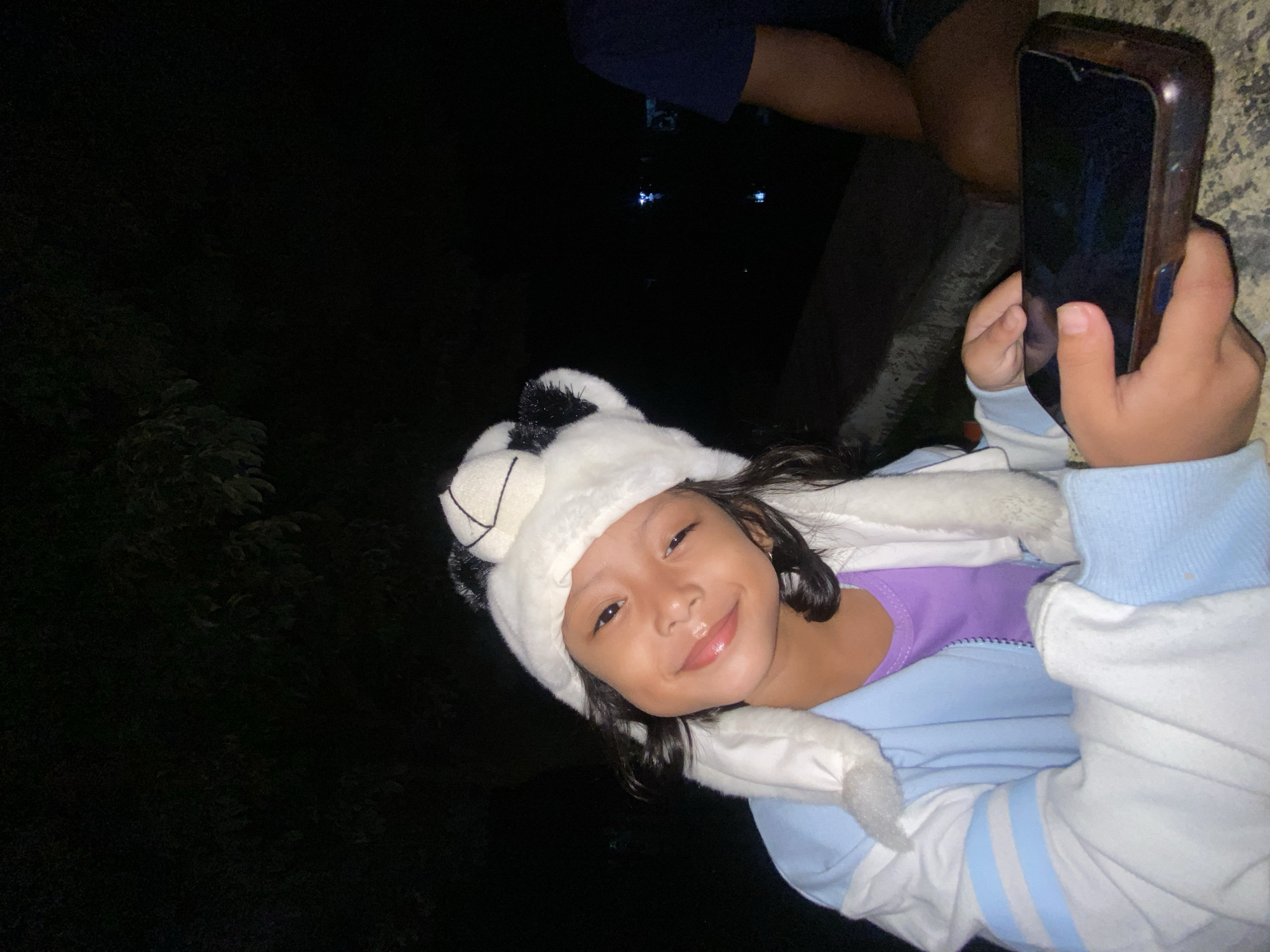 A young girl is holding a smartphone, possibly taking a selfie. She is wearing a white and black panda hat. She is also wearing a light blue jacket over a purple top. The background is dark, possibly outdoors at night. Some dark trees are visible in the background. Parts of two other people appear in the image. The photograph was taken in Kalitengah, Indonesia.