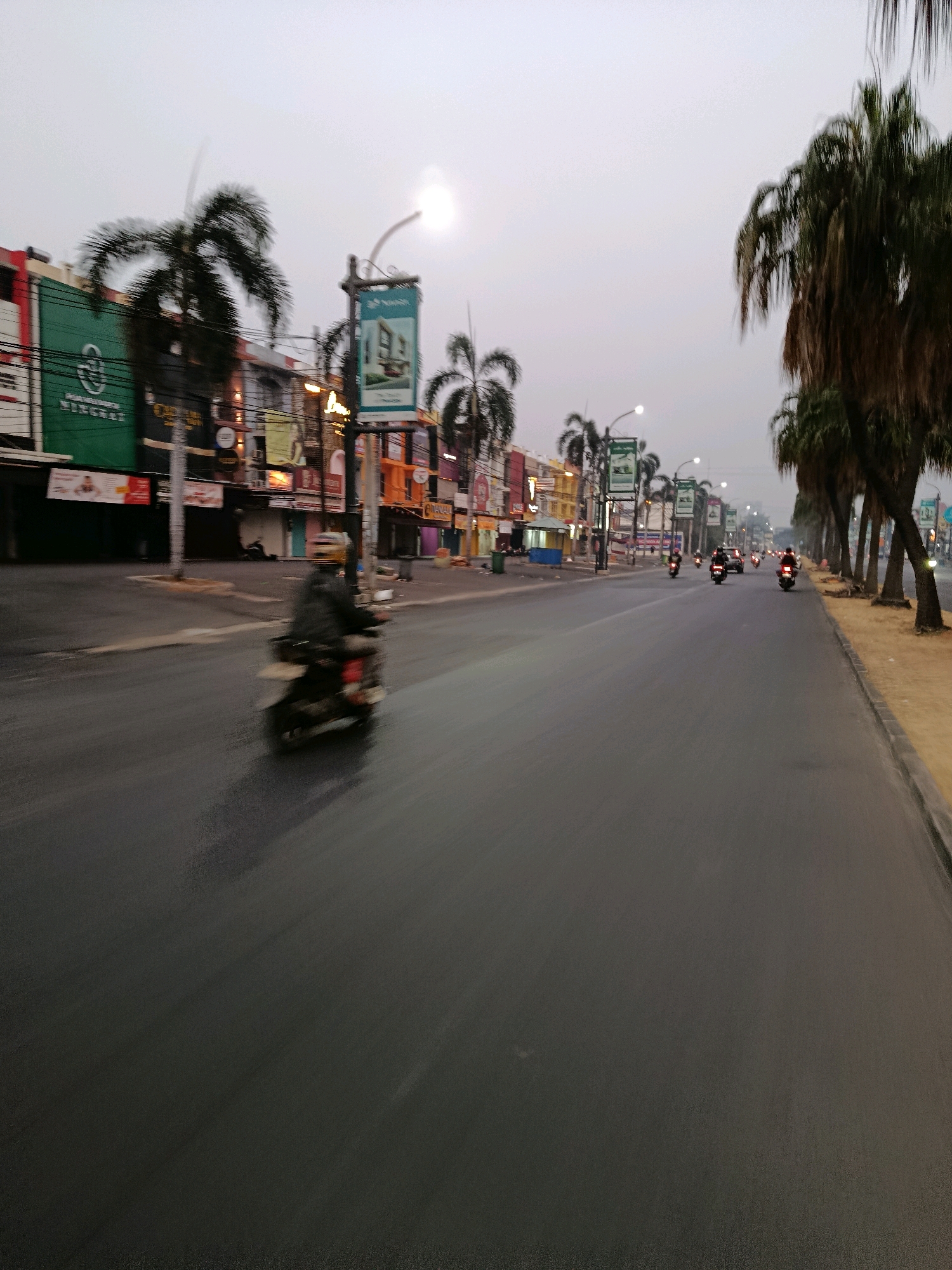 This is a medium-shot, eye-level, outdoor, action photograph taken in a city street of Bekasi, Indonesia. The image depicts a street scene during a cloudy day, likely early morning or late afternoon, with overcast skies. The wet asphalt road reflects the muted light. On the left side of the road, there is a row of buildings with various signs and advertisements. Palm trees line the right side of the road.
In the foreground, a person on a motorcycle is moving away from the camera. The motorcycle and rider are blurred due to motion. Further down the street, several other motorcycles and a car are visible, also in motion. The street appears to be relatively empty of pedestrian traffic, suggesting a quiet time of day. Streetlights are present along the road. The overall impression is a typical urban street in Bekasi, with a focus on the movement of vehicles.