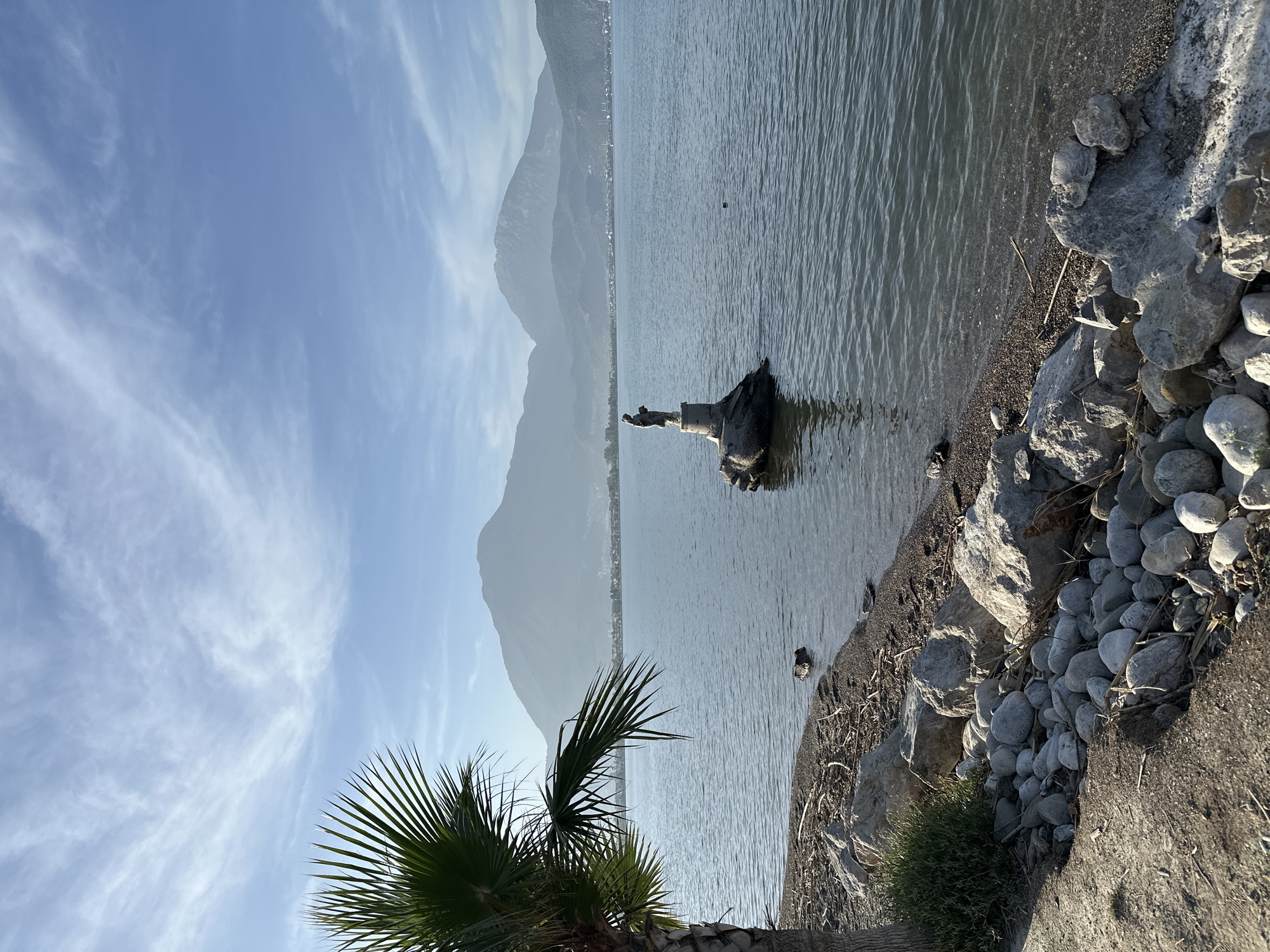 The image shows a body of water next to a rocky shoreline with a statue of a person on a pedestal in the water. The background features mountains and a clear sky with wispy clouds. A palm tree is visible in the foreground. The statue is situated close to the shore, partially submerged. The scene appears to be in Fethiye, Turkey.