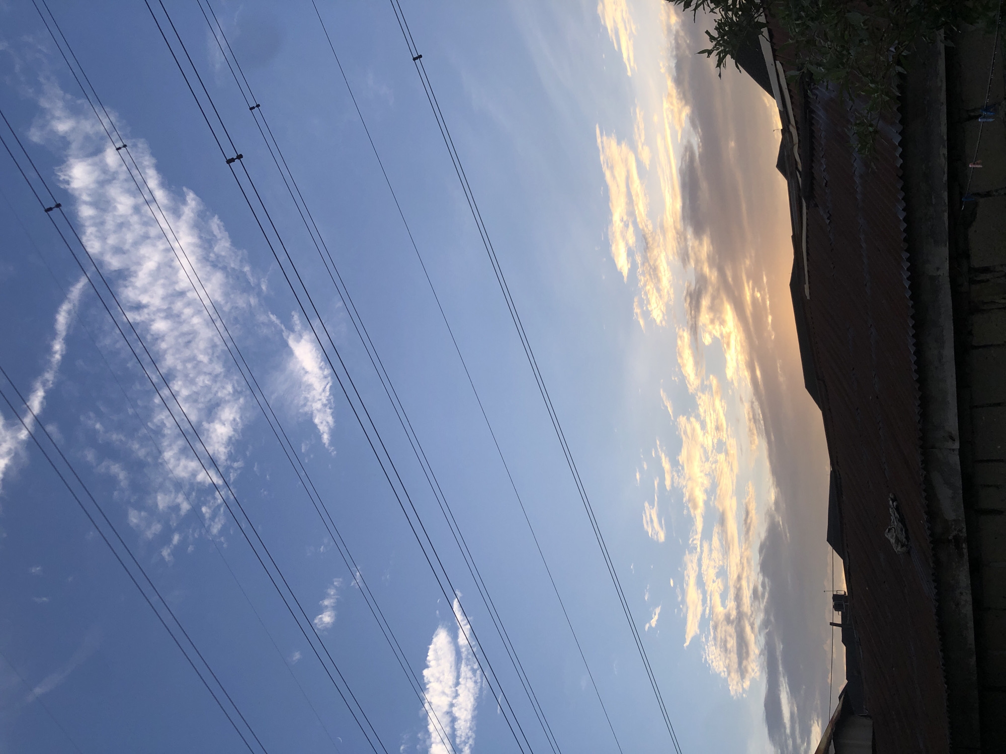 The image depicts a view of a cloudy sky with sunset colors over rooftops in Ado, Nigeria. Several parallel power lines stretch across the frame, diagonally from the upper left to the lower right. The sky transitions from a deep blue at the top to lighter shades of blue and orange, reflecting the setting sun, with scattered white and grey clouds. Below the sky, the corrugated metal roofs of several buildings are visible, appearing somewhat weathered. Some greenery from trees is present on the right side of the frame, and a small section of a concrete wall is at the bottom. There are no people or animals clearly visible in the image.