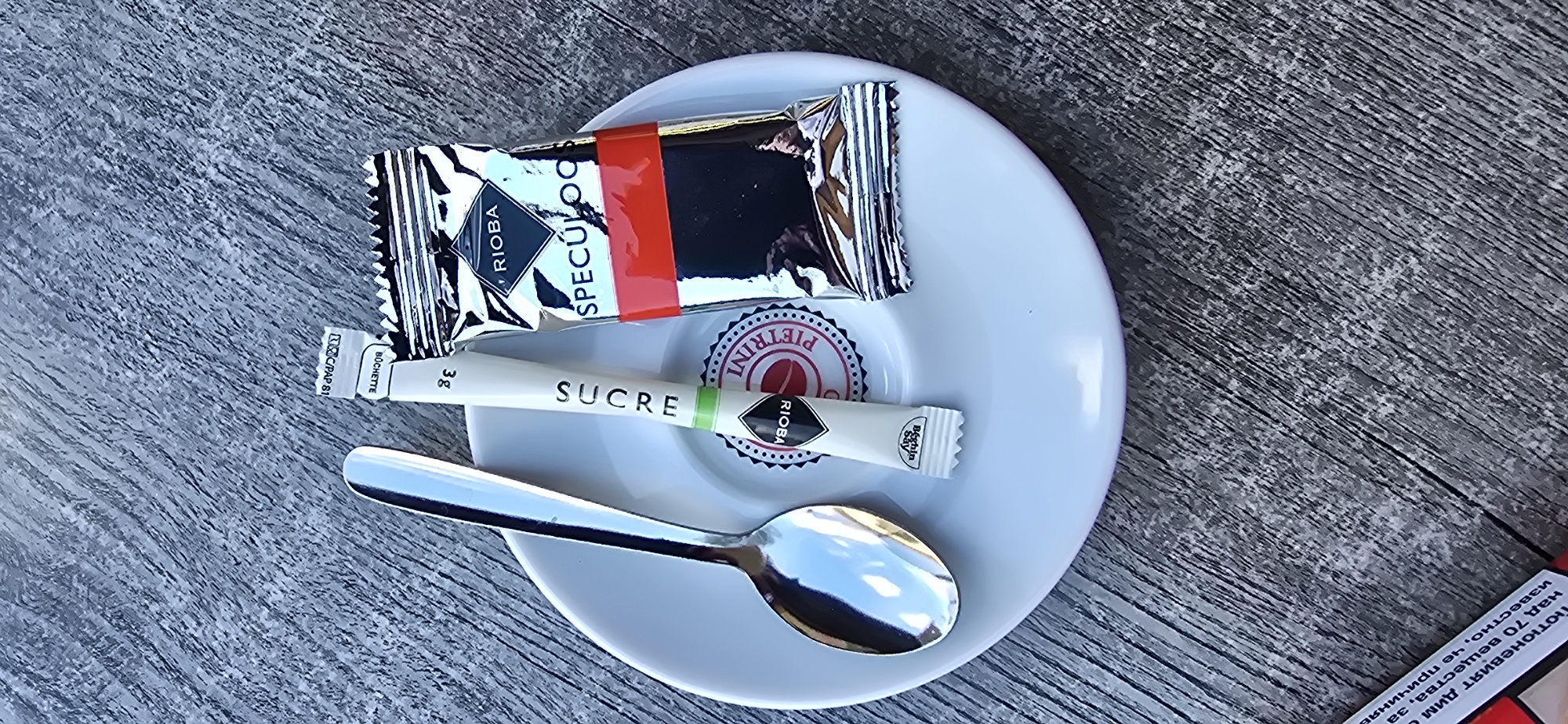 A close-up overhead shot captures a white saucer on a gray wooden table. On the saucer are a silver spoon, a white sugar packet labeled "SUCRE", and a silver packet of "RIBA SPECULOOS" cookies. The sugar packet is topped with a small white label. A circular logo with the words "PIETRINI RIOBA" is visible on the saucer beneath the sugar packet.