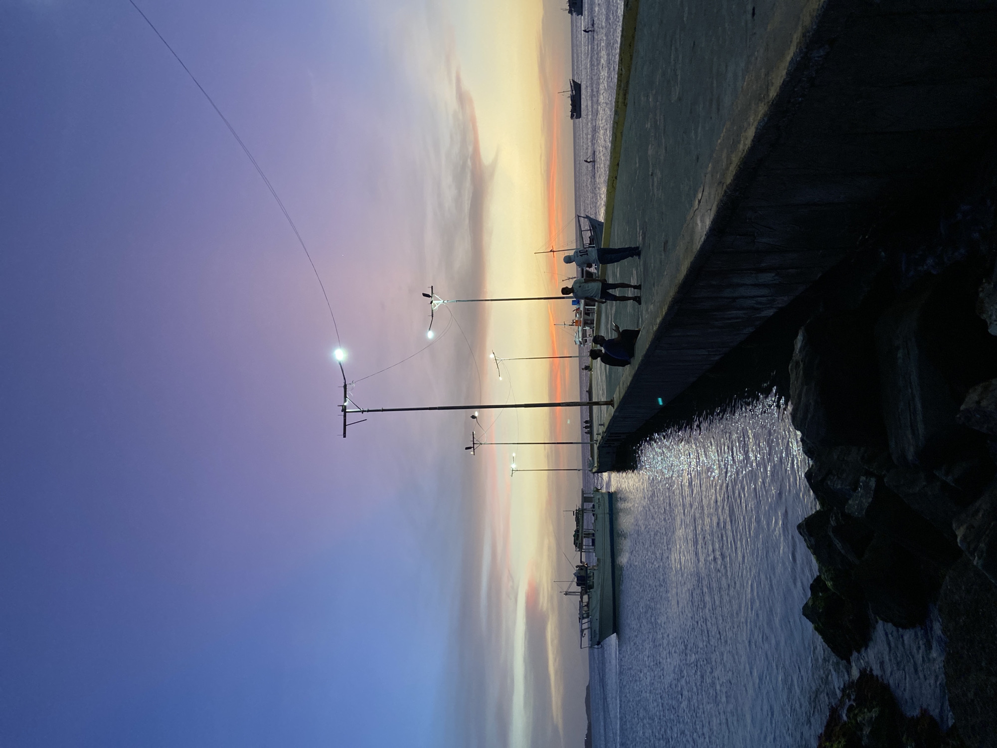 Here's a description of the image:
The image depicts a waterfront scene at dusk in Juan Griego, Venezuela. The sky transitions from blue to orange and yellow near the horizon, with some cloud cover.
A concrete walkway runs along the water's edge. Several people are standing on the walkway. There are light posts lined up along the walkway. The water is somewhat reflective.
Boats are visible in the distance, both in the water and docked nearby. Rocks and possibly a seawall are present at the edge of the water in the foreground.