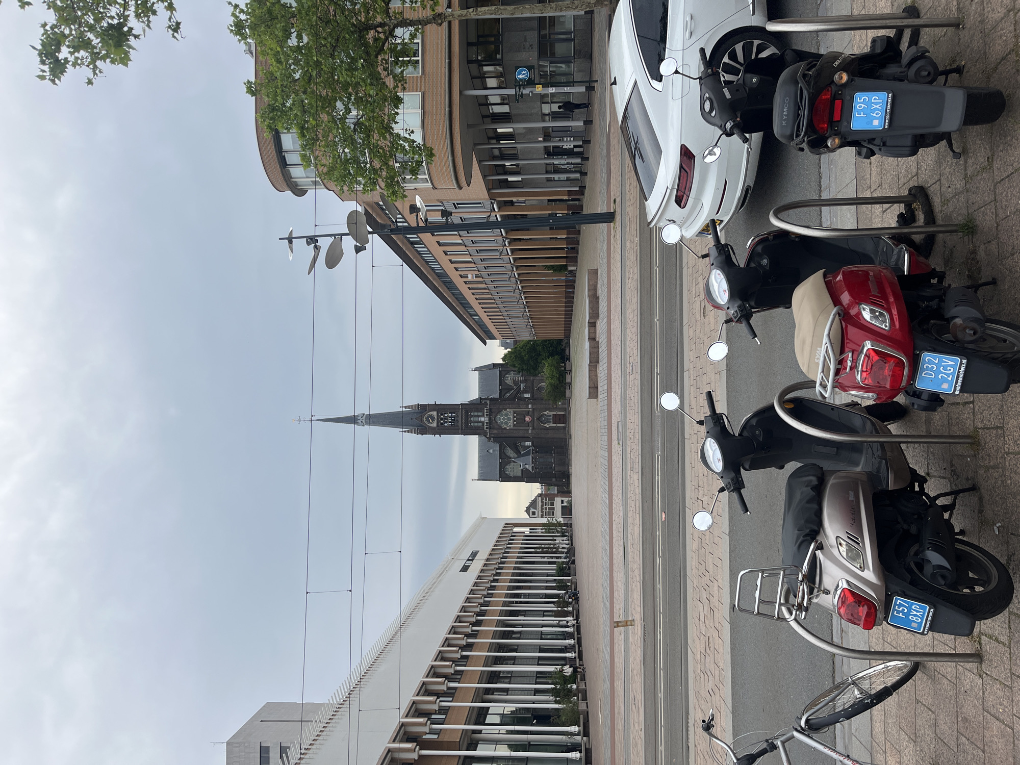 A wide, overcast shot captures a European city square. In the foreground, three scooters are parked, one beige, one red, and one black. A silver bicycle is parked to the left of the scooters. Behind the scooters, a white car is parked. In the background, a large, dark-colored church with a tall spire dominates the center of the frame. To the left of the church is a long, modern building with a colonnade, and to the right, a curved brick building. The sky is cloudy, and the overall lighting is diffused.