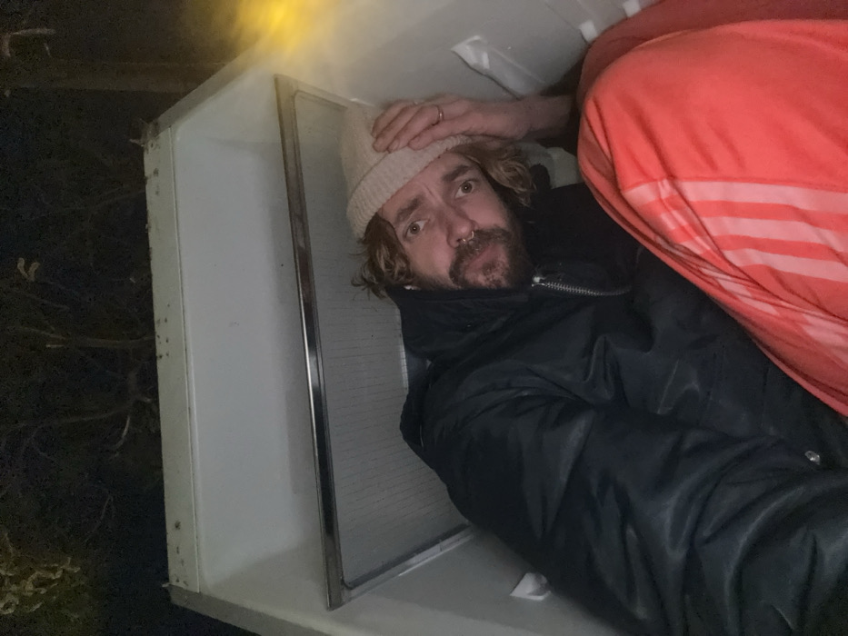 A person with a tan beanie, curly brown hair, and a black jacket lies inside a white refrigerator. Their head is near the top left corner with a hand on their head, while an orange and white striped garment is visible on the right. The refrigerator stands outdoors, suggested by the dark background featuring foliage. The person gazes into the camera with a serious expression.