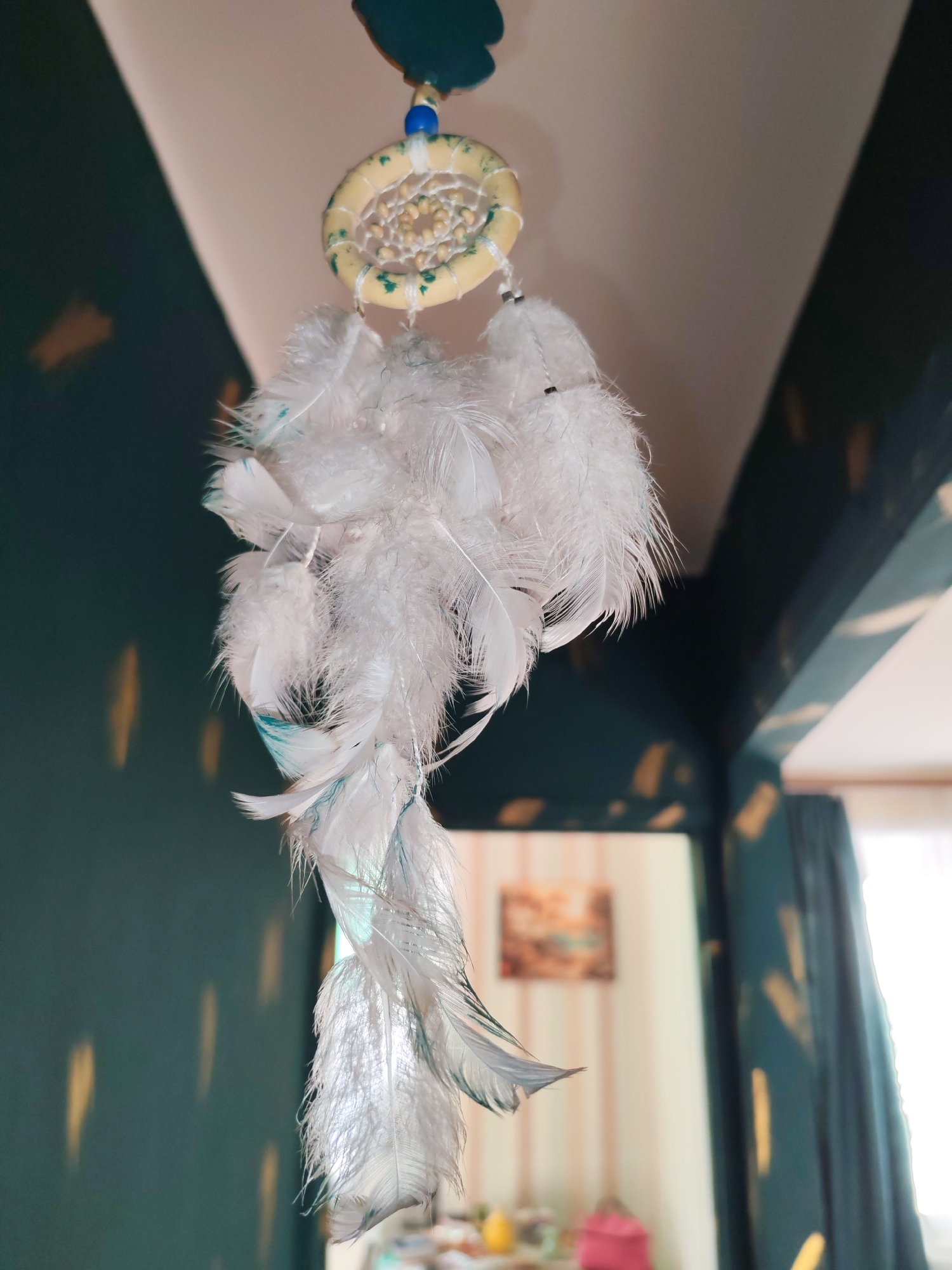 A dreamcatcher hangs suspended in an indoor setting. The dreamcatcher features a circular frame with an intricate web inside, adorned with white feathers. The upper portion of the dreamcatcher has a blue bead and a dark green decorative piece. The background shows a room with dark green patterned walls, a doorway leading to another room with light-colored walls and an indistinct picture, and curtains partially obscuring a window. The location or city of Izhevsk, Russia is not evident in the image.