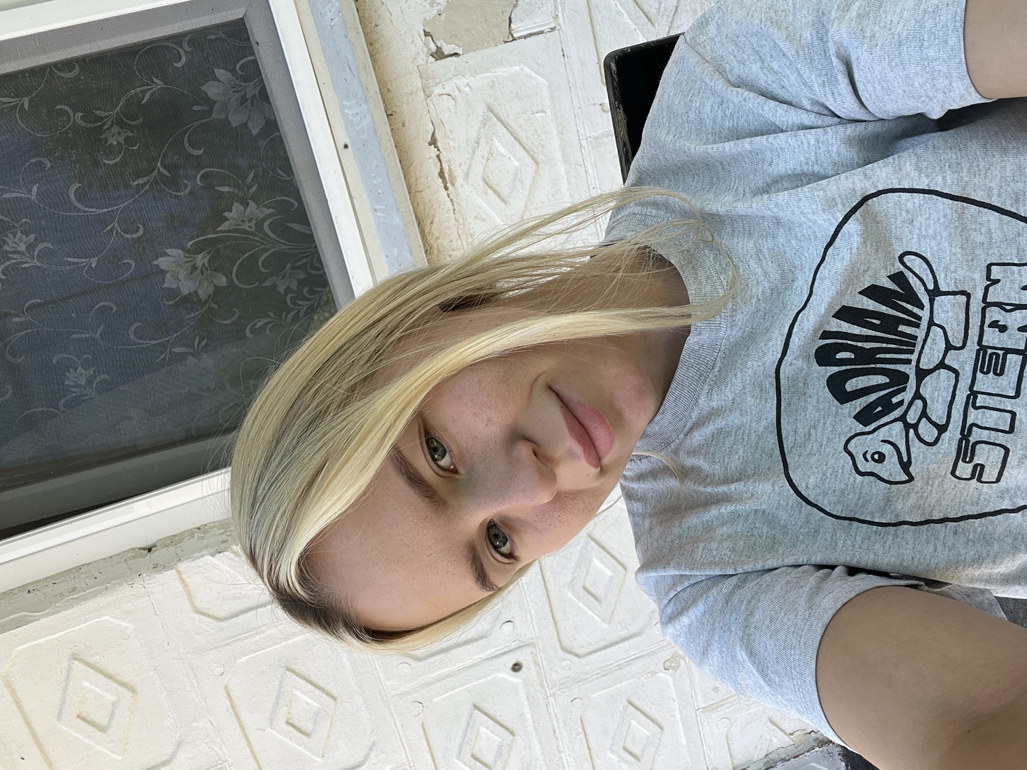 A young woman with blonde hair is captured in a selfie, her face angled slightly towards the camera. She is wearing a grey t-shirt with a black graphic design of a cartoon turtle and text. The background consists of a white brick wall with a textured diamond pattern and a window with a floral screen. The lighting suggests an outdoor setting during the daytime. The image does not provide any information about the specific location within a city.