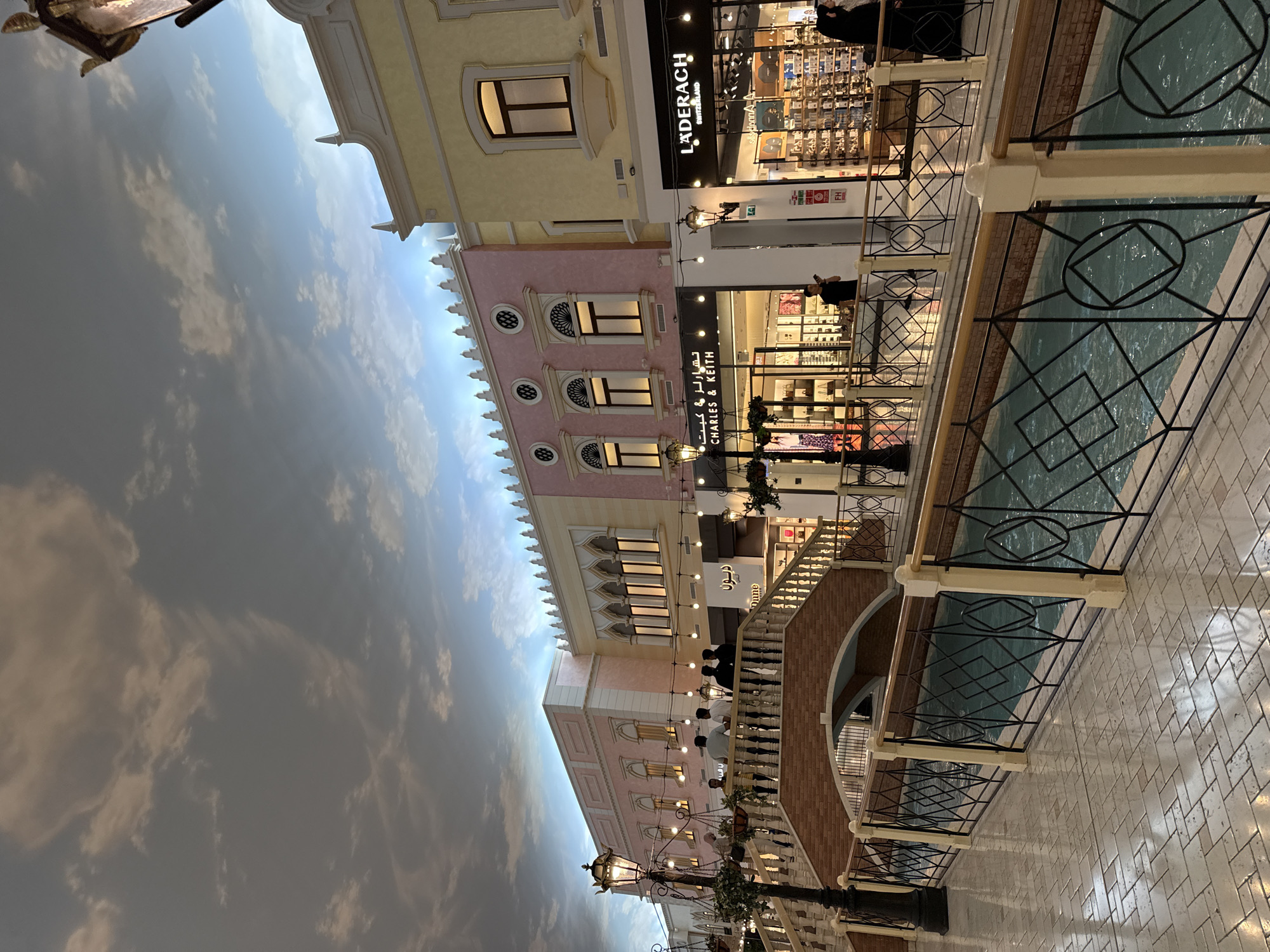 The image shows the Villaggio Mall in Al Rayyan, Qatar, featuring an indoor canal with buildings resembling Venetian architecture. The "sky" above is painted with clouds. Several shops are visible, including Ladurach and Charles & Keith. People are present along the canal walkways. A decorative bridge arches over the canal. The walkway is paved with light-colored stone. Balustrades line the canal. There are lampposts with hanging plants visible as well.