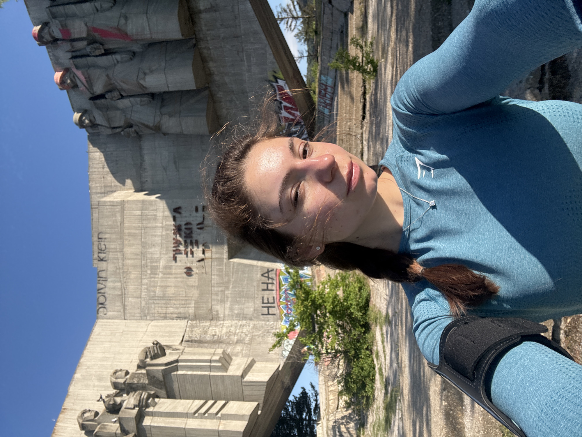 A woman is posing for a selfie in front of the Buzludzha Monument in Bulgaria. The monument, which is a large concrete structure with statues and graffiti, is situated on a hill overlooking the surrounding landscape. The woman is wearing a blue shirt and has her hair in a braid. She is holding her phone in her right hand. In the background, the sky is blue.