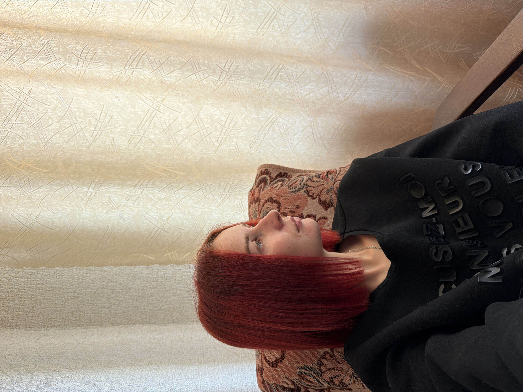 A woman with red hair lies on a patterned armchair. She is wearing a black sweatshirt with raised black lettering. The background consists of textured wallpaper. There is no information in the image to determine the location is Chaikovsky, Russia.