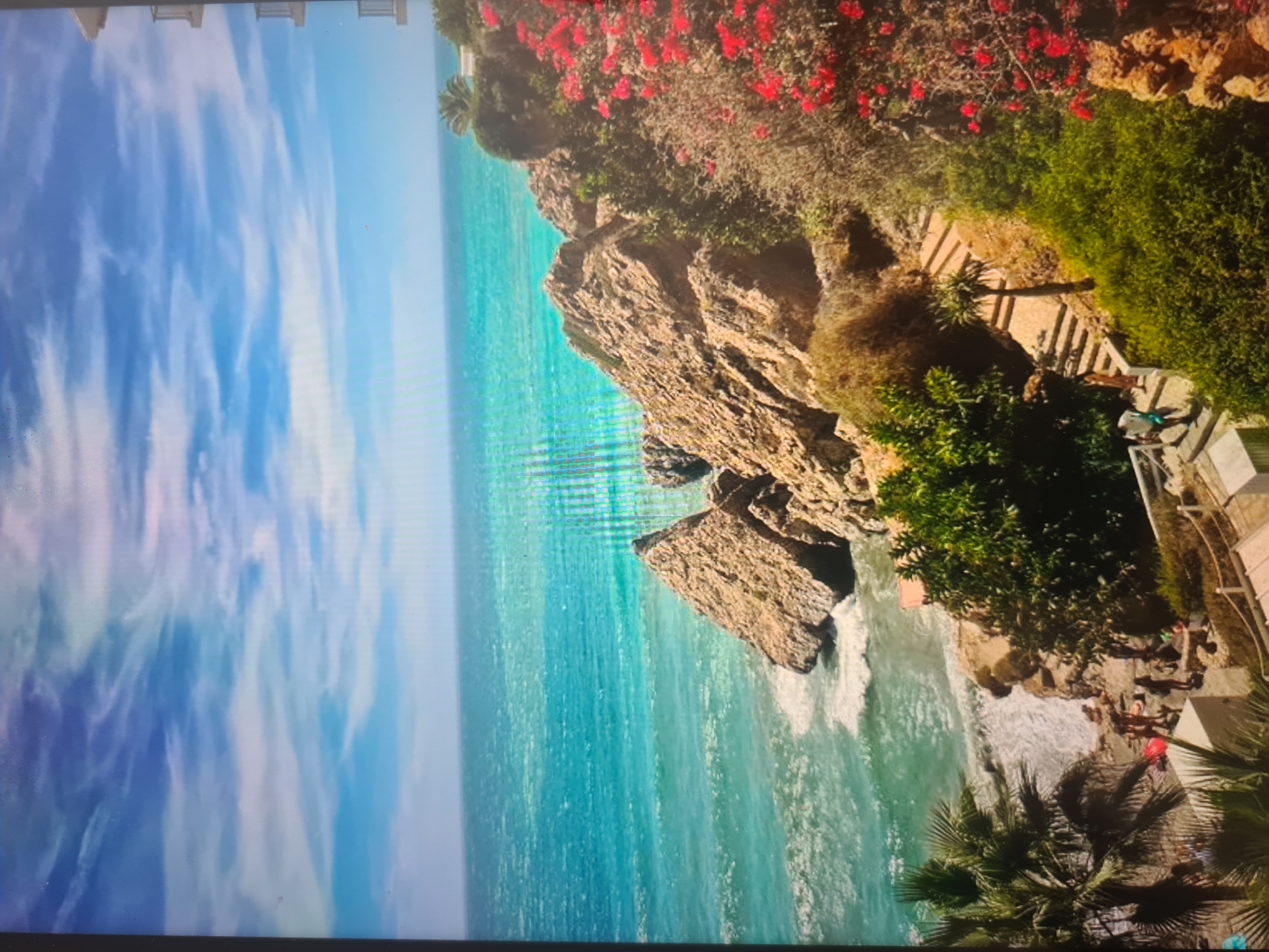 The image, when viewed with the sky at the top, displays a vibrant coastal landscape under a bright, partly cloudy sky. The vast expanse of the sky is a brilliant blue, adorned with wispy white clouds. Below, a vivid turquoise-blue sea stretches towards a rugged and dramatic coastline.
The prominent feature of the land is a series of rocky cliffs and outcroppings that meet the water. These cliffs are densely covered with lush green vegetation, including several palm trees and bushes with striking red flowers, possibly bougainvillea, adding splashes of color. A paved path or steps, likely carved into the terrain, descends along the right side of the image, leading down to a small, secluded beach or rocky cove.
Several people are visible in the lower right portion of the scene. Some figures are relaxing or standing near the water on the sandy/rocky shore, while one person appears to be walking on the steps. A red object, possibly a towel or blanket, is visible on the beach. Waves gently break against the shore and the base of the cliffs, creating white foam. Part of a building or structure with a light-colored wall can be seen on the far right edge.
The scene depicts a beautiful and serene coastal destination, likely enjoying sunny weather during the day. The clear visibility and vibrant colors suggest a pleasant atmosphere, possibly in a Mediterranean or tropical region. No visible text is present in the image.