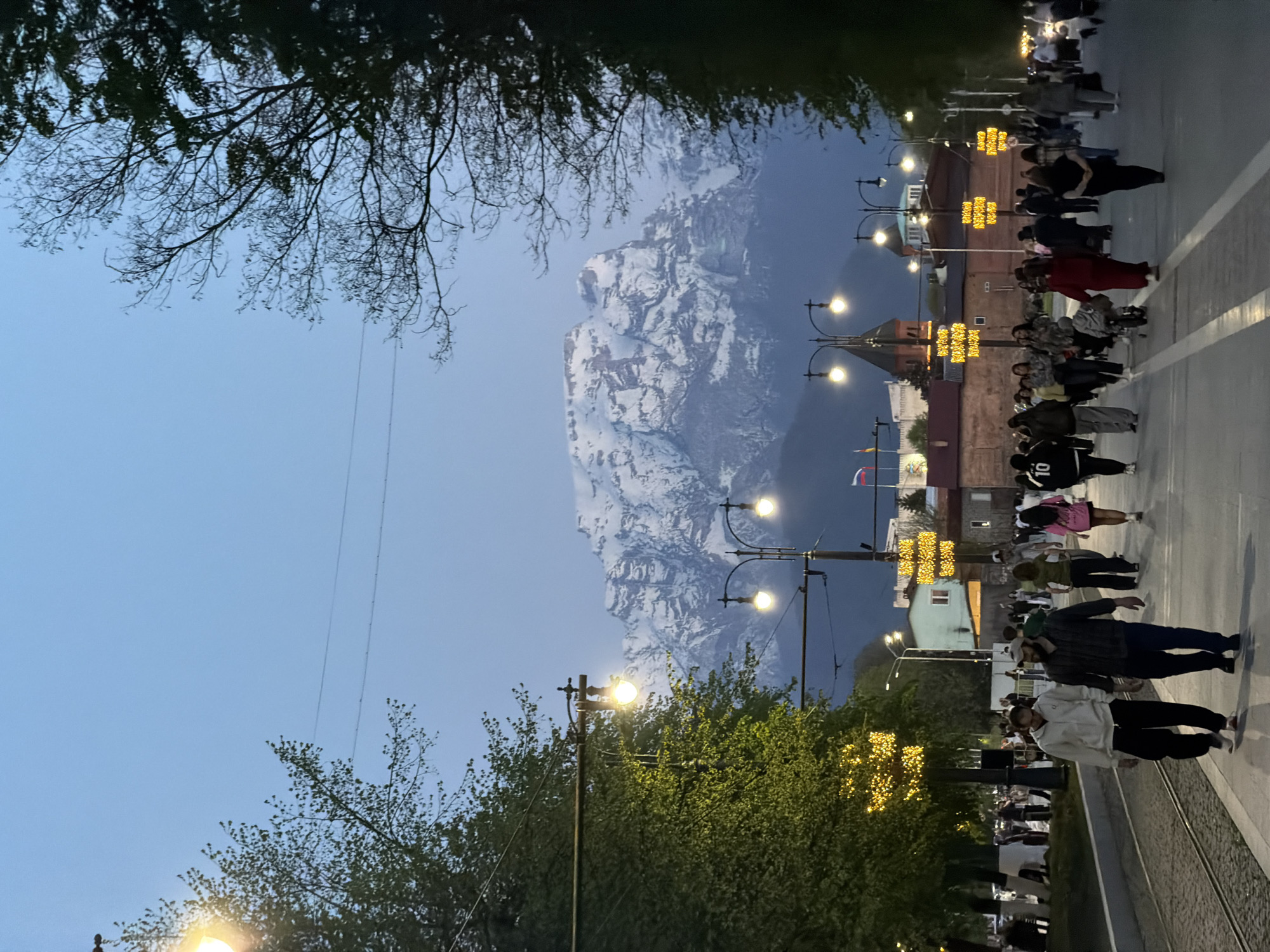The image presents a street scene in Chermen, Russia, at dusk. A snow-capped mountain forms a backdrop in the distance. A row of illuminated lampposts lines the street. People are walking on the sidewalk along the tramway track. Trees flank the street. Some branches are bare while others are covered in leaves. The sky is light blue. A Russian flag is visible near the buildings.