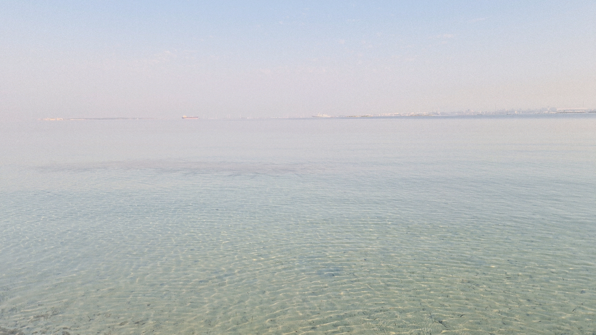 The image shows a calm, shallow body of water with a sandy bottom visible through the clear water. The horizon is visible in the distance, where a hazy city skyline can be seen. A few distant ships are also present on the water. The sky is a pale blue and appears to be overcast or experiencing haze. There are no people or animals visible in the image. The scene suggests a tranquil coastal environment. Given the description of a hazy city skyline and ships, this could be a view of the Mar Piccolo or Mar Grande in Taranto, Italy, as both bodies of water are adjacent to the city and are part of its port.