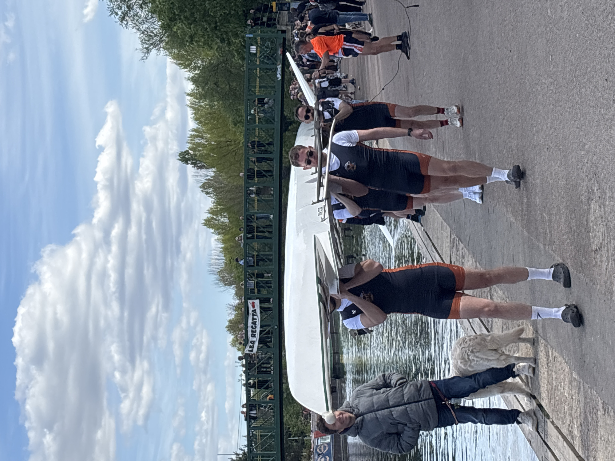 A partially obstructed, slightly tilted long shot captures a scene at the Lea Regatta in London, United Kingdom. The focus is on a rowing team consisting of four individuals in black and orange sportswear, carrying a white rowing boat. They are accompanied by a man in a gray jacket and jeans, as well as a white dog. A man in an orange shirt stands further back. In the background, a bridge with spectators overlooks the river. The sky is blue with scattered clouds.