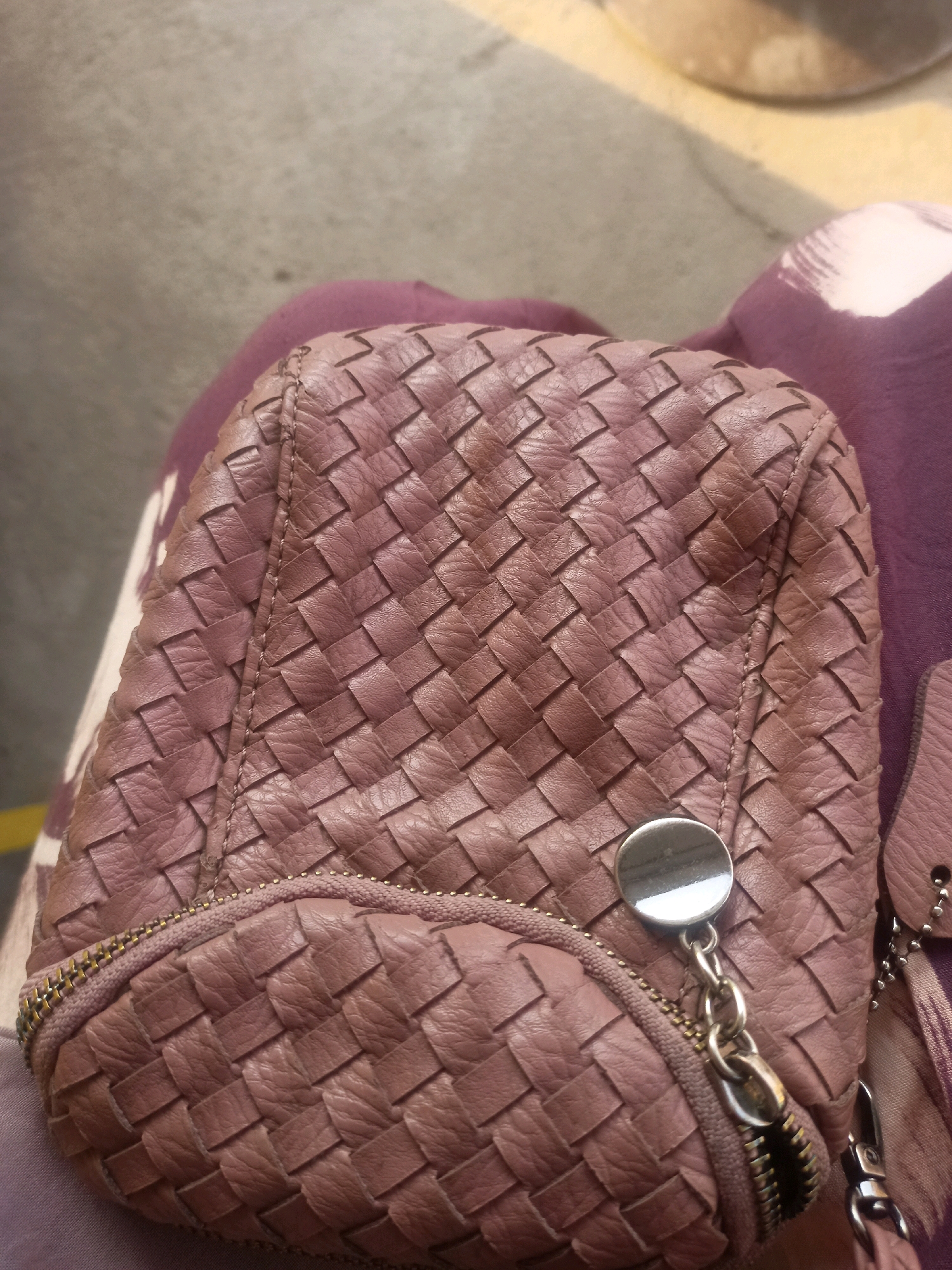 The image shows a close-up view of a woven, pinkish-brown leather clutch bag. The bag has a textured, braided pattern. A silver circular clasp is attached to the front of the bag by a chain with multiple links. A zipper with gold-toned teeth is visible along the bottom edge of the bag. The bag is resting on a piece of purple fabric, which appears to be part of clothing. In the background, a neutral-colored, textured surface, possibly a floor or pavement, is visible. The image does not provide any information about a specific location in Ketro, Indonesia.