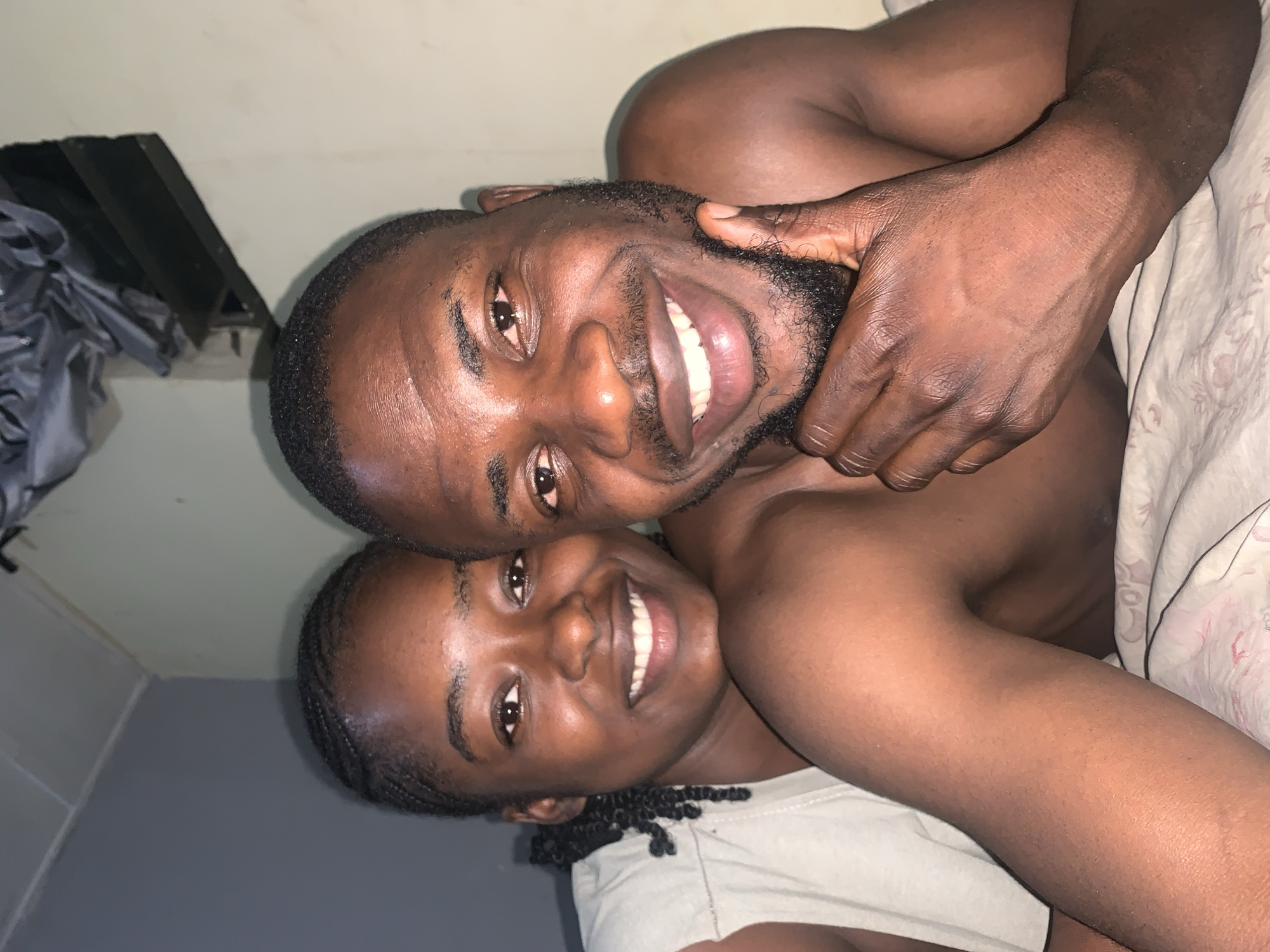 This intimate, close-up portrait shows two smiling individuals, likely a couple, embracing and looking directly at the camera. The image is composed with the subjects appearing as if they are lying down and the camera is angled above them.
The man, positioned towards the top and right of the frame, is shirtless, showcasing his dark skin tone and muscular physique. He has a short, dark beard and neatly cropped hair. His left arm is gently wrapped around the woman's head, his hand resting affectionately near her cheek. He displays a wide, joyful smile, with his eyes reflecting clear happiness.
Nestled close to him, the woman is seen slightly below and to the left. She wears a light-colored top and has dark, braided hair. Her face mirrors the man's, illuminated by a broad, radiant smile as she looks towards the viewer. Both individuals exude a sense of warmth, happiness, and affection for each other.
The background is minimal, featuring a plain, light-colored wall (possibly off-white or light grey) that fills most of the space. In the lower left, a darker grey surface is visible. A light-colored blanket or sheet with a subtle pattern can be seen on the right side, suggesting an indoor, cozy setting, possibly a bed. Some dark, possibly crumpled fabrics or objects are visible in the top left corner. The lighting is soft and warm, highlighting their expressions. The scene evokes a feeling of comfort and connection, taking place in Rafin Par, Nigeria. There is no visible text in the image.