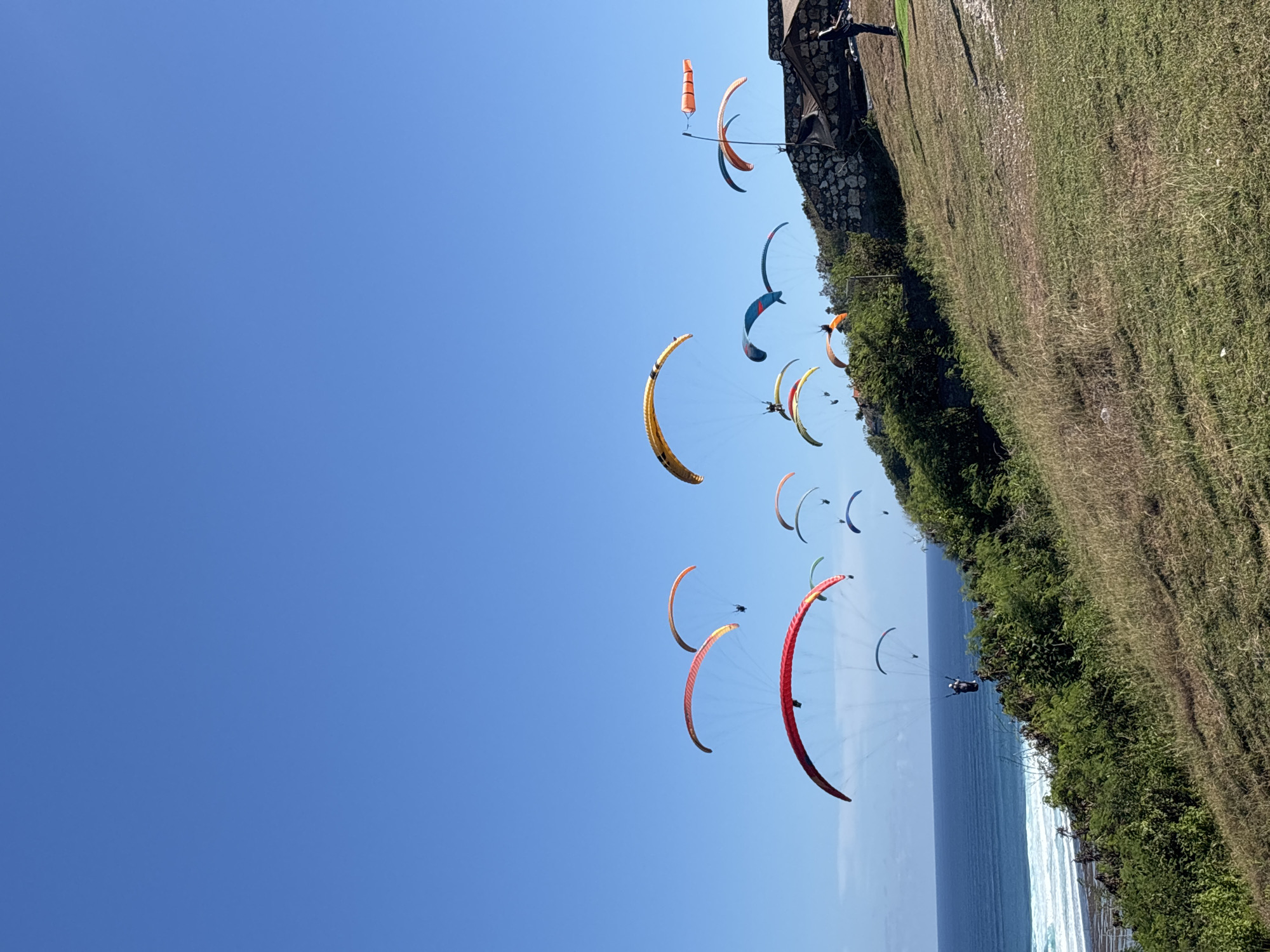 The image shows a group of paragliders soaring in the sky above a cliff overlooking the ocean. The sky is clear and blue. The cliff is covered with green vegetation. There are numerous paragliders with various colors and designs visible. The location is Kuta Selatan, Indonesia, as evidenced by the distinctive coastal terrain and paragliding activity common in the area.