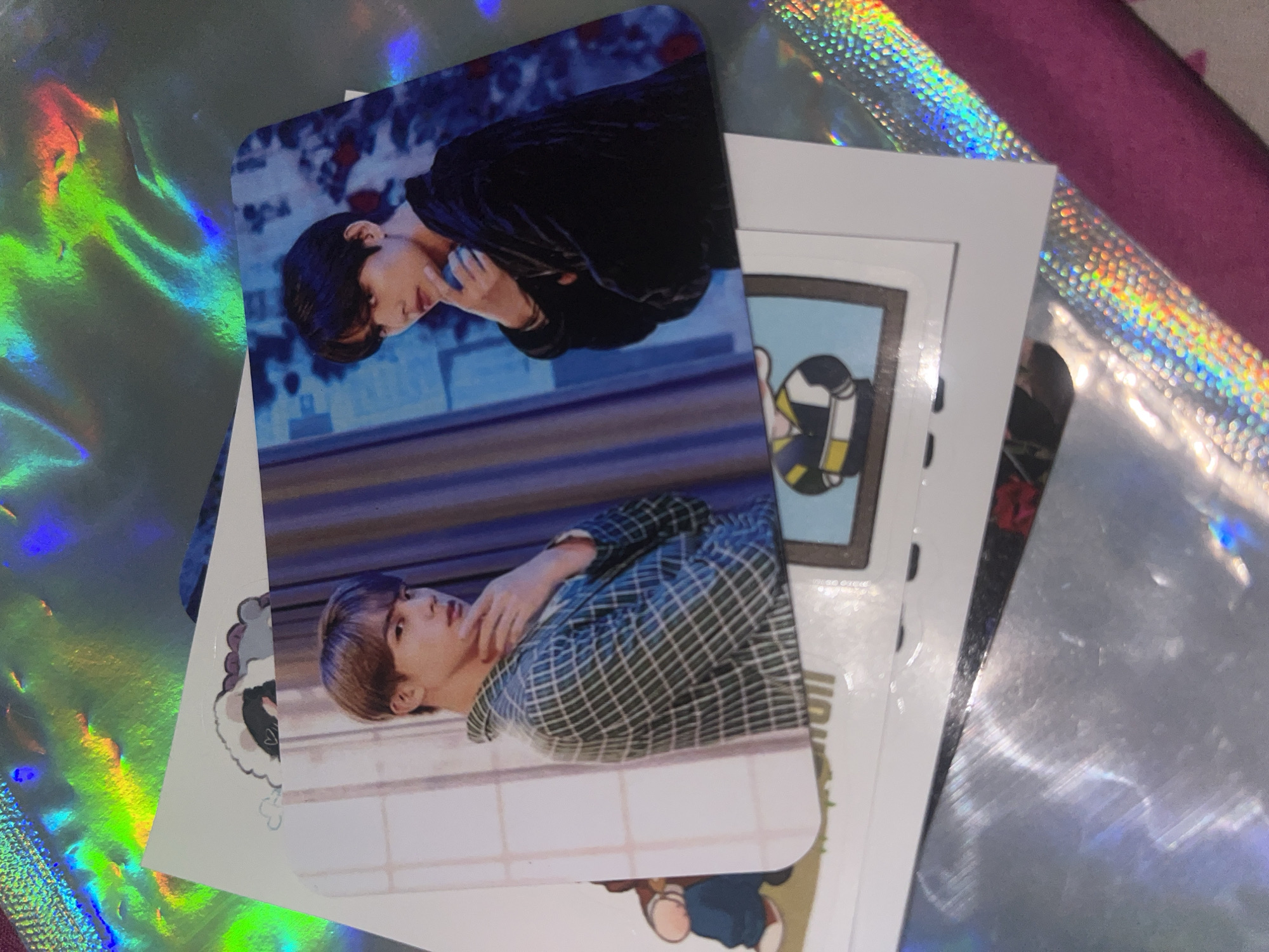 The image shows a stack of cards with different images on them. The topmost card features a portrait of a man with short brown hair, wearing a green and white plaid shirt, and resting his hand near his neck. The card is positioned horizontally. Partially overlapping it is another card with a different portrait of a man, wearing a black velvet jacket with a blue background. The other cards in the stack beneath these two have various cartoon-like illustrations. The background shows a shiny, iridescent surface, along with a pink surface. The location in the city of Central Jakarta, Indonesia is not evident.