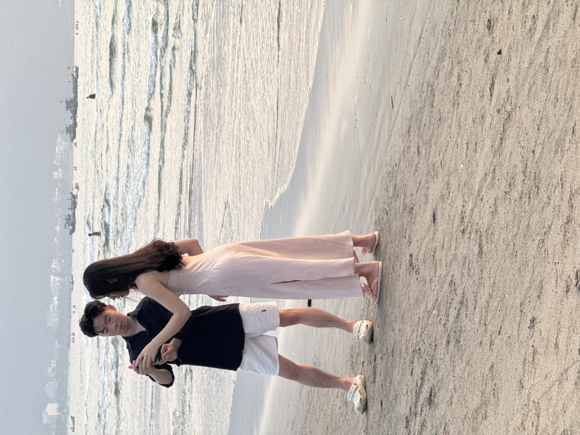 A full shot depicts a couple standing on a beach in Đà Nẵng, Vietnam. The man, in a dark navy shirt and white shorts, is holding a small electronic device, possibly a camera or phone, in his hands, appearing to examine or adjust it. The woman, wearing a light pink dress, stands close to him, her head tilted down as if looking at the device. Both are wearing casual beach footwear.
The scene is set on a sandy beach, with the ocean waves visible in the background. In the distance, the city’s skyline can be seen, obscured by a light haze or mist. The beach appears relatively empty, with no other individuals visible nearby. The photo’s aspect ratio and composition suggest it may have been taken on a phone. The lighting indicates it is either early morning or late afternoon.
