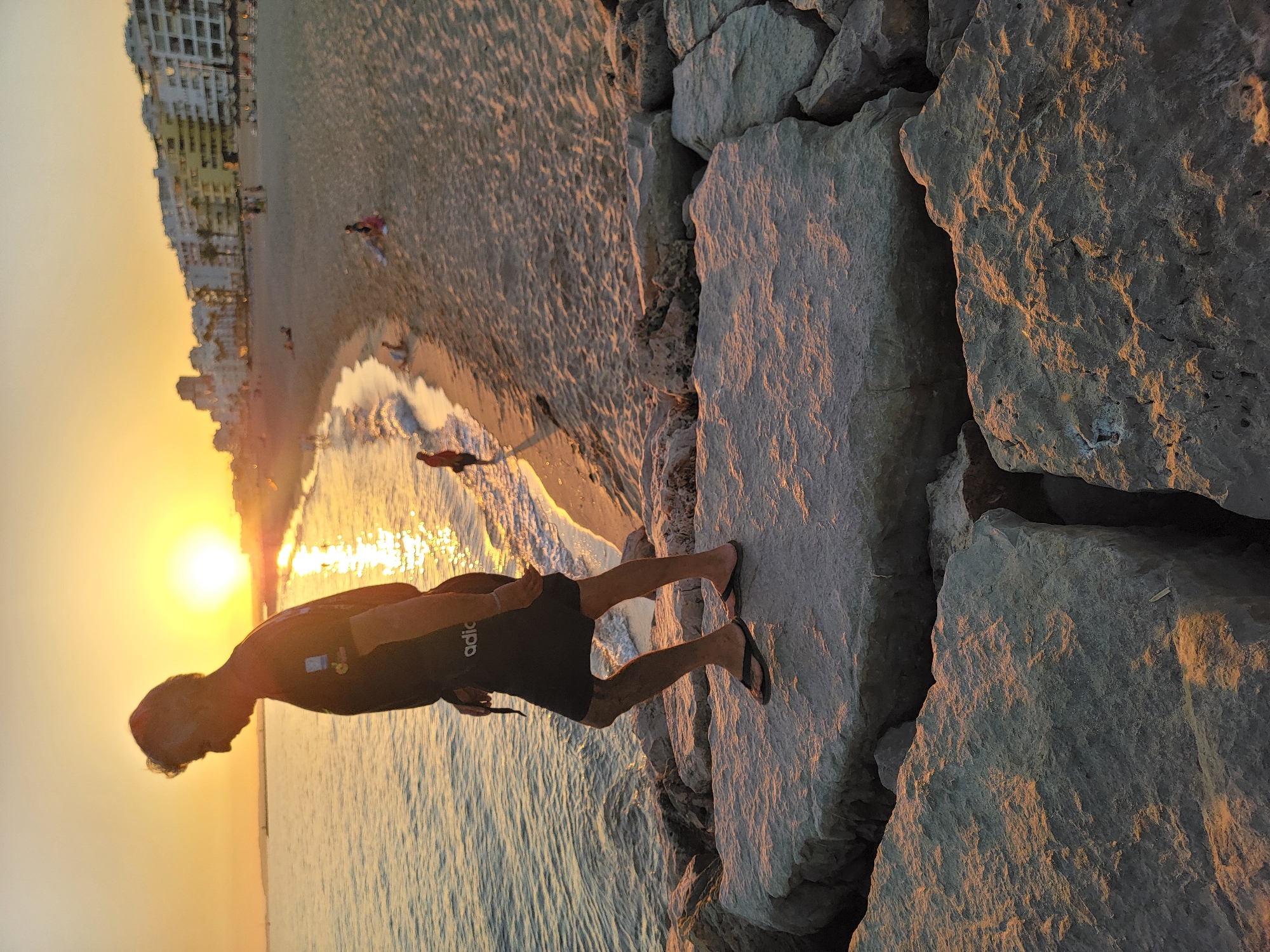 An elderly man wearing a black t-shirt and black shorts with flip-flops is walking on large, flat rocks at the edge of the water. The sun is setting, casting a warm orange glow over the scene. Behind him, a sandy beach stretches out with several buildings and palm trees. People are scattered on the beach and in the water. The location is Quarteira, Portugal.