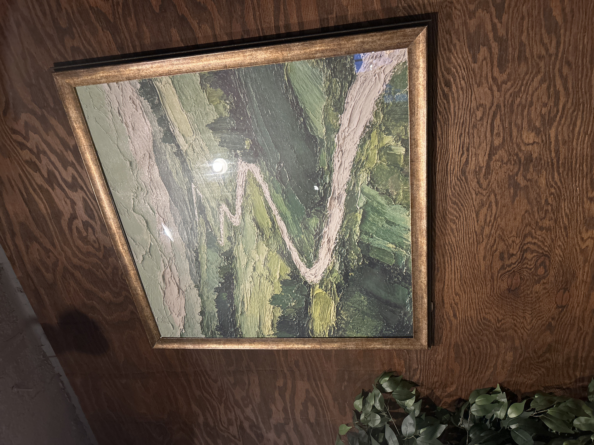 A painting of a winding road through green hills is displayed on a wood-paneled wall. The painting has a thick, impasto style with visible brushstrokes. A golden frame surrounds the artwork. To the left of the painting, a branch of an artificial plant with green leaves is visible. The wood paneling has a prominent grain pattern. There is a shadow cast on the wall above the painting, suggesting a light source from the right. The overall setting appears to be indoors.