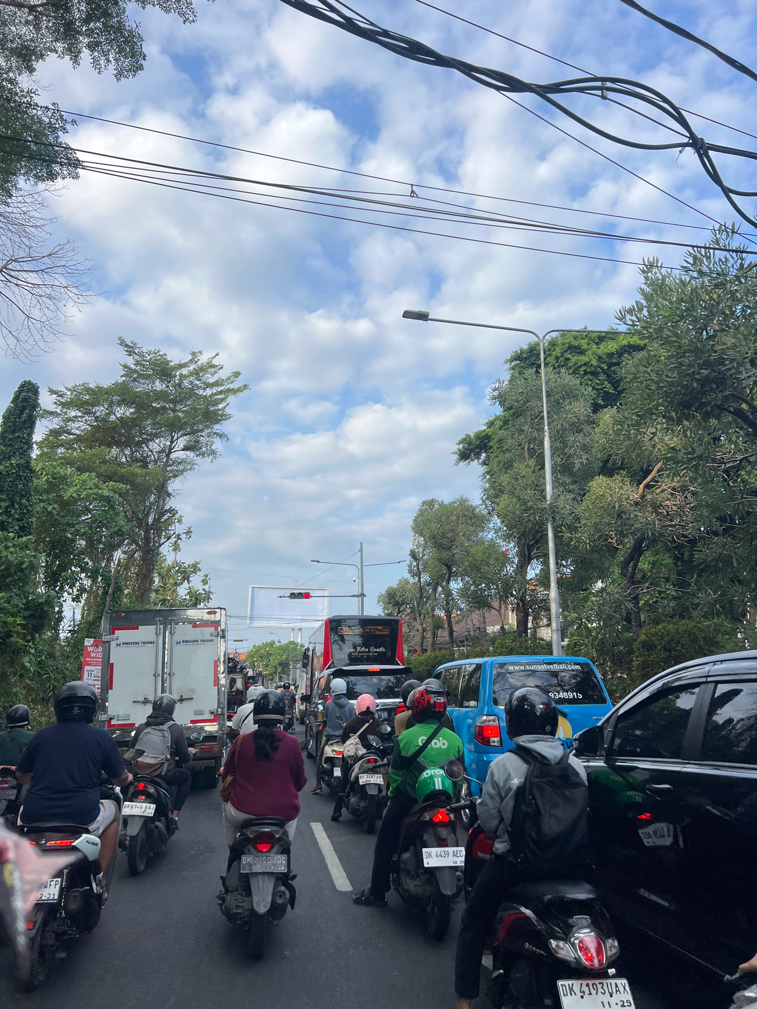 The image captures a congested traffic scene on a street in Denpasar, Indonesia. A multi-lane road is filled with numerous motorcycles and a few cars and trucks. People on motorcycles are wearing helmets, suggesting adherence to traffic regulations. The vehicles are stopped, indicating a traffic jam. Trees and foliage line the sides of the road, with overhead power lines and streetlights visible. The sky is partly cloudy, with a bright, diffused light. The overall impression is a typical urban traffic situation in a tropical climate.