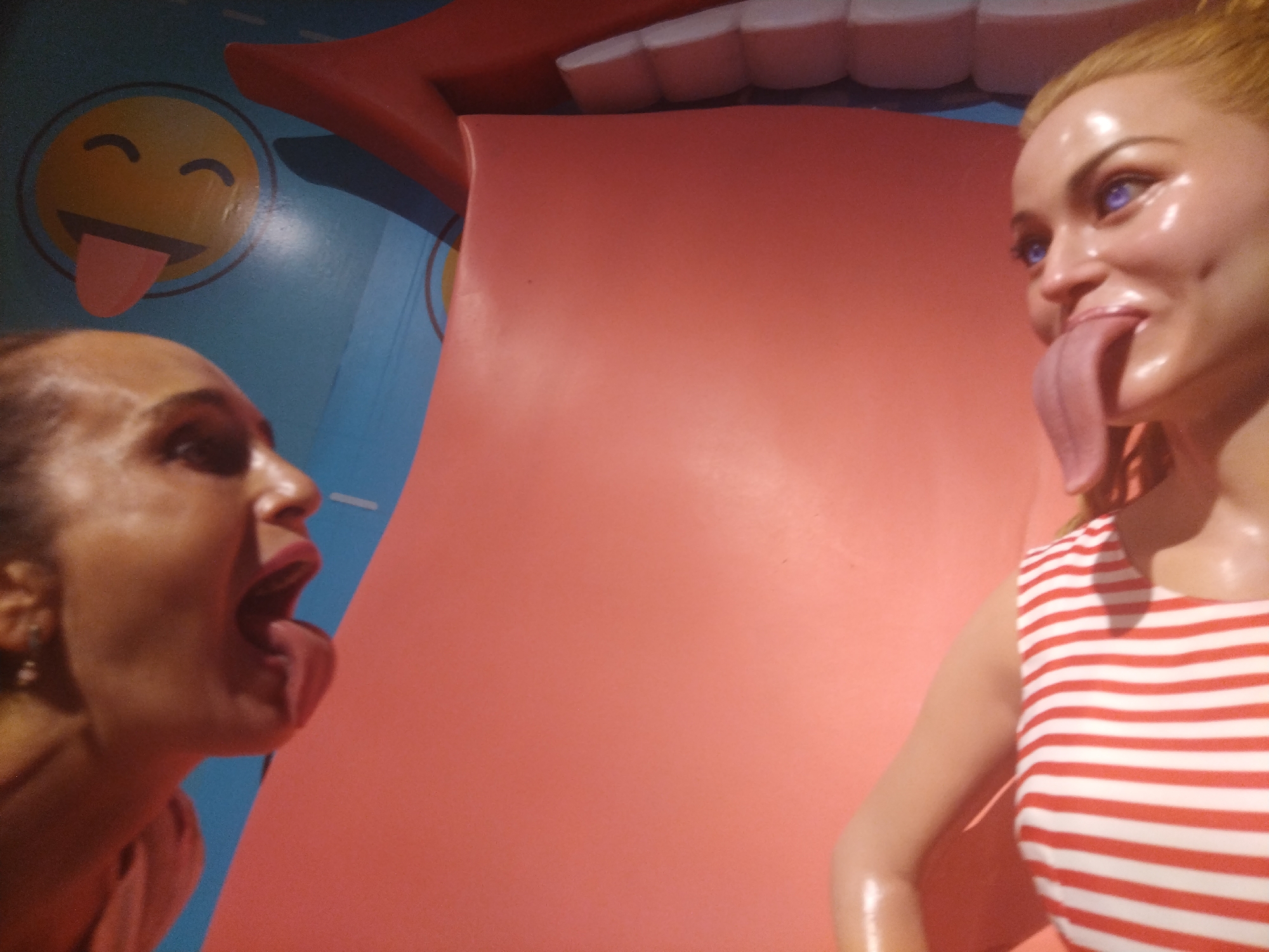 The photograph captures a woman and a mannequin engaged in a playful interaction at what appears to be a tourist attraction in the city of Unknown, Unknown. The woman is positioned on the left side of the frame, with her mouth open and tongue extended towards the mannequin. The mannequin, dressed in a striped red and white top, reciprocates the action with its own extended tongue. Above them, a large, stylized representation of a mouth with white teeth is visible. In the background, a blue surface features a yellow emoji sticking out its tongue. The setting suggests a lighthearted, possibly humorous, environment.
