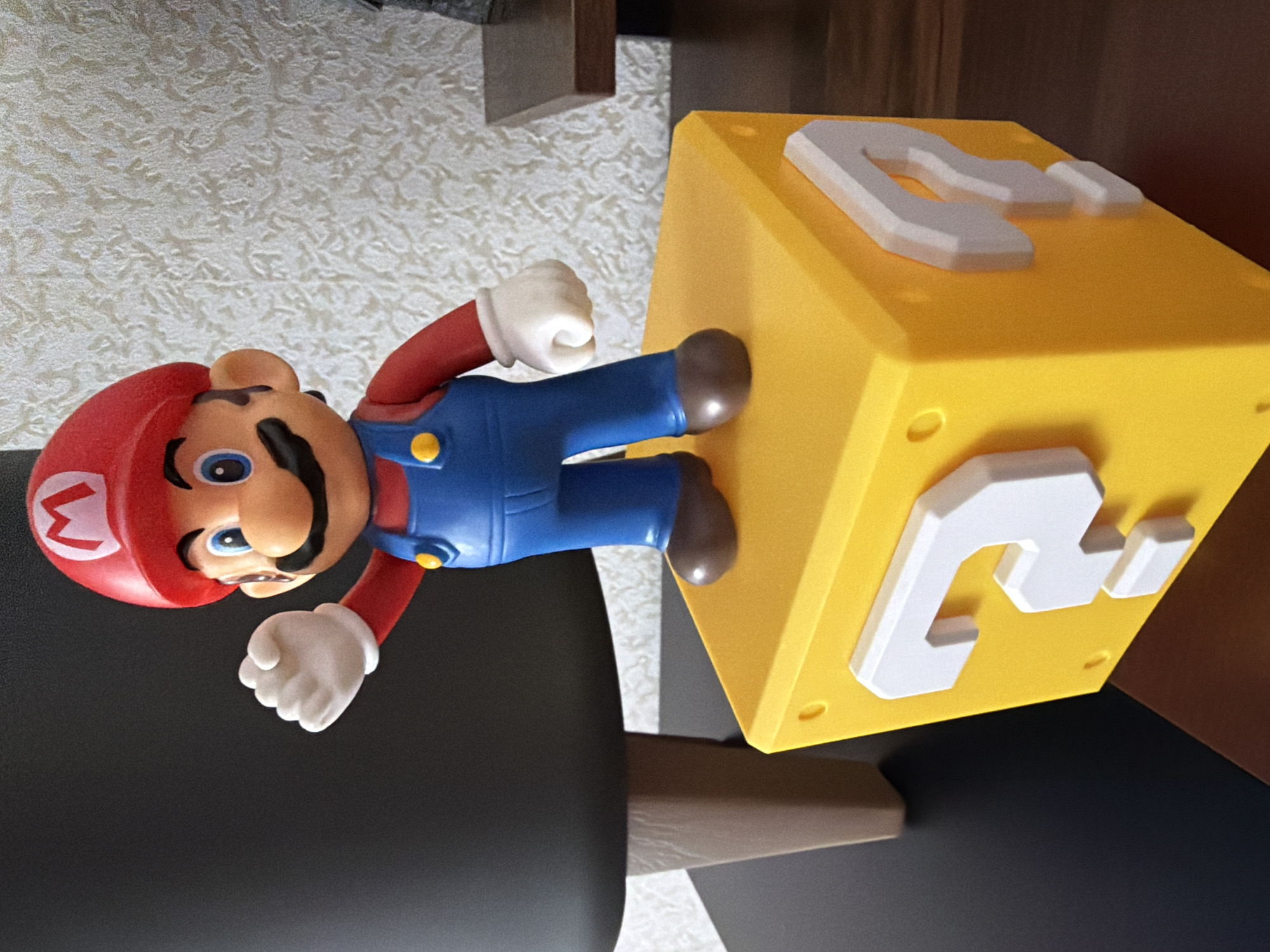 A detailed forensic, matter-of-fact description of the image:
The image shows a Mario figurine standing on top of a yellow, cube-shaped object with question marks on its sides. The figurine is of the character Mario from the Nintendo franchise, wearing his signature red hat with an "M" logo, a red shirt, blue overalls, and brown shoes. He has white gloves and is positioned with his right arm raised and hand open, as if waving. The cube has raised white question marks on its visible faces. The scene is lit by overhead light, casting soft shadows. The background consists of a dark chair on the left, a textured, off-white wall behind the Mario figurine, and a wooden surface with a dark brown hue on the right, possibly a desk or shelf, on which a dark object, possibly a speaker or a decorative item, is partially visible. The overall setting suggests an indoor environment, likely a home or office. The precise location within the city of Yablonovskiy, Russia, cannot be determined from the visual information provided.