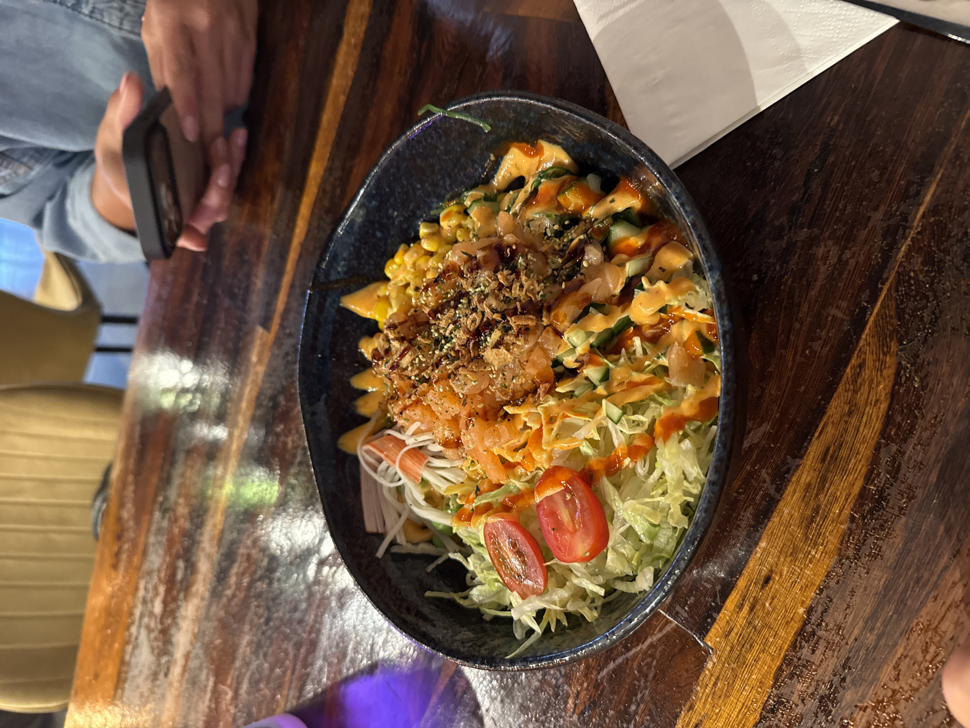 In Groningen, Netherlands, a poke bowl is presented on a dark wooden table. The bowl is dark blue and filled with a variety of ingredients including shredded lettuce, sliced tomatoes, cucumber, corn, imitation crab sticks, and chopped salmon. A creamy orange dressing is drizzled over the top, along with crispy fried onions and sesame seeds. A person's hands, wearing a light blue denim jacket, are visible in the background, holding a smartphone. The background also shows a yellow chair with vertical stripes. The lighting suggests an indoor setting, possibly a restaurant.