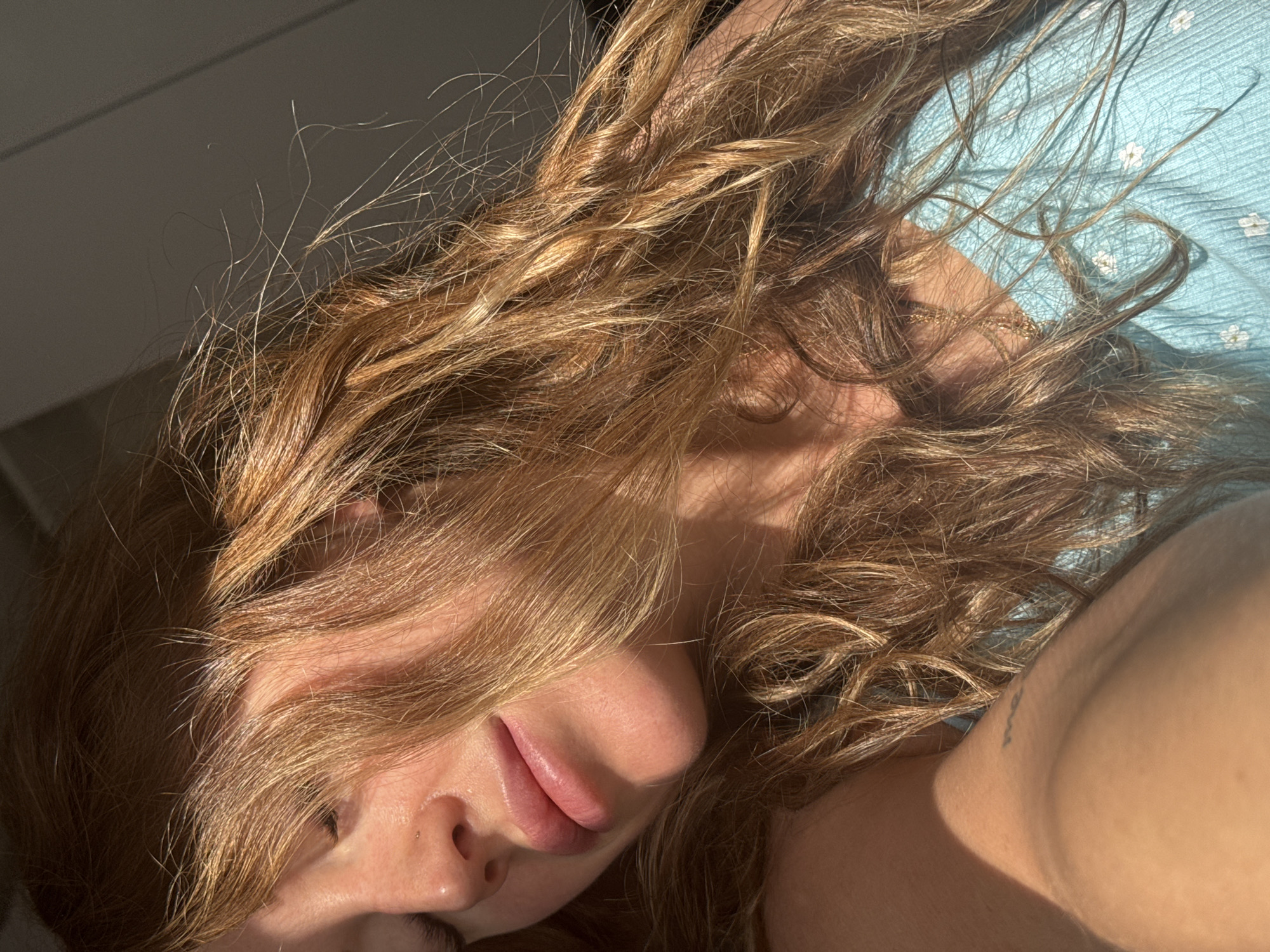 A close-up, tilted, medium shot captures a woman with long, wavy brown hair partially obscuring her face. Her eyes are closed, and her lips are slightly parted. She appears to be lying down. Her bare shoulder and part of a blue garment with a floral pattern are visible. There is a tattoo on her shoulder. The lighting is harsh, with strong contrasts of light and shadow. The location is not discernible from the image. The image does not provide information about actions, interactions or the environment.
