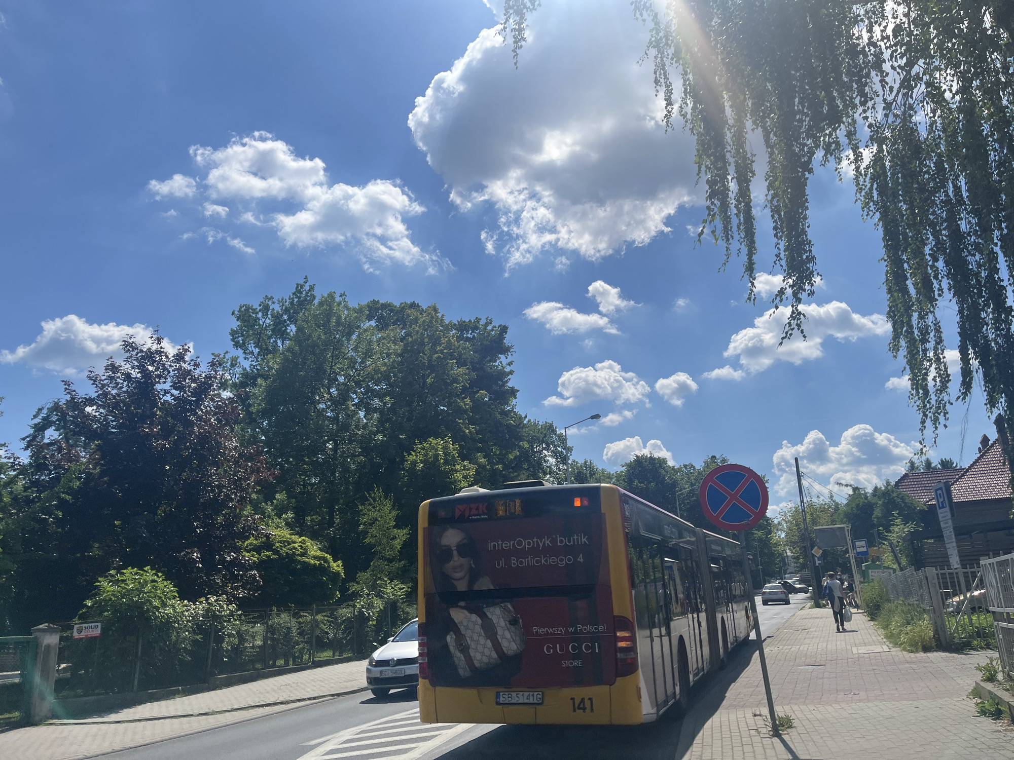 This is a wide-angle, eye-level shot of a street scene under a bright, sunny sky with scattered white clouds. The sun is high, suggesting it's midday. Lush green trees line the street, with some deciduous trees exhibiting dark red foliage and others displaying their full summer green. On the right side of the road, buildings with red-tiled roofs are visible.
The main subject is a yellow public bus with advertisements on its side. One advertisement prominently features the Gucci logo and a woman wearing sunglasses and carrying Gucci bags, along with text in Polish that translates to "First in Poland! Gucci Store." The bus also displays the route number "141" and a license plate reading "SB 5141G." A white car is visible driving in the opposite direction on the left side of the road.
Further down the street, a pedestrian walks away from the camera on a paved sidewalk. A blue and red circular sign with a red cross inside indicates a "No Stopping" zone. Another sign further down the sidewalk appears to be a bus stop sign. The overall atmosphere is calm and casual, characteristic of a typical summer day in a suburban or urban residential area. The sunlight casts shadows and creates lens flare in the upper right portion of the image, indicating a bright and clear day.