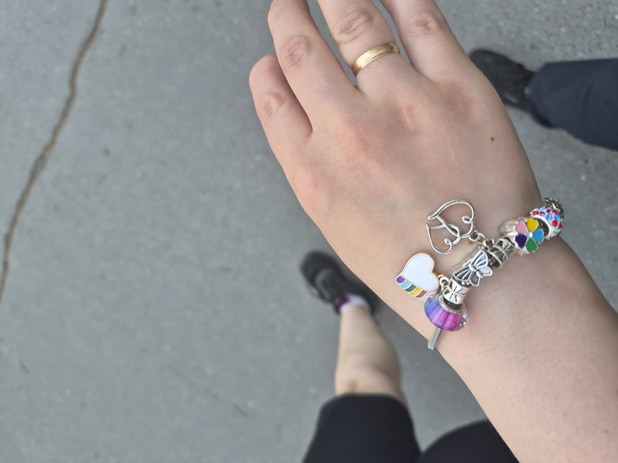 A fair-skinned person, likely female based on the jewelry and visible clothing, is standing on what appears to be a grey sidewalk or paved surface. The person is wearing a gold wedding band on their left ring finger and a charm bracelet on their right wrist. The bracelet includes a heart charm, a butterfly charm, and other colorful decorative beads. Parts of a leg wearing a black shoe and a leg wearing a black short, and a black shoe, are visible in the lower portion of the image, and part of a navy blue pant leg with black shoe is visible on the right, suggesting the person is walking or standing with at least one other individual. There is no evidence to indicate that this picture was taken in the city of Tobolsk, Russia.