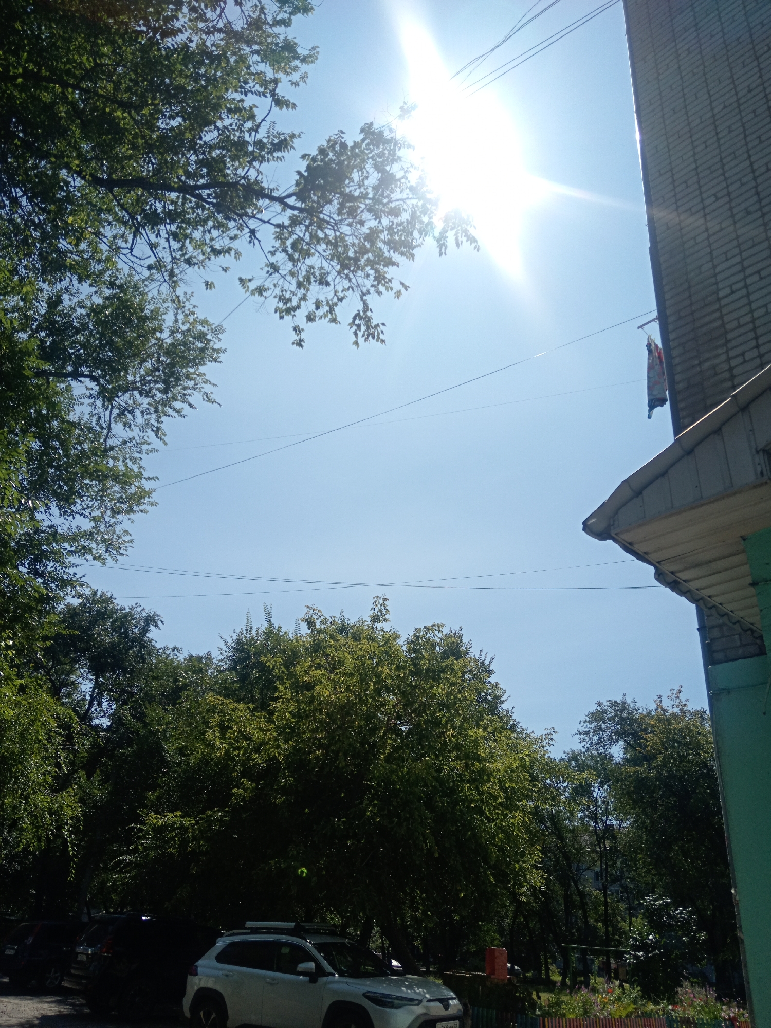 The image shows a bright, sunny day in Belogorsk, Russia. The sky is clear blue with the sun shining intensely, creating lens flare. On the right side of the frame, a brick building with a gray overhang and a green vertical section is visible. A clothesline with laundry hangs on the side of the building. Lush green trees with dense foliage fill the lower and left portions of the image, with branches extending towards the sky. In the foreground, several parked cars are visible, including a white SUV and darker vehicles. The scene appears to be an outdoor residential area, likely a courtyard or street in front of apartment buildings, given the presence of the building, trees, and cars.