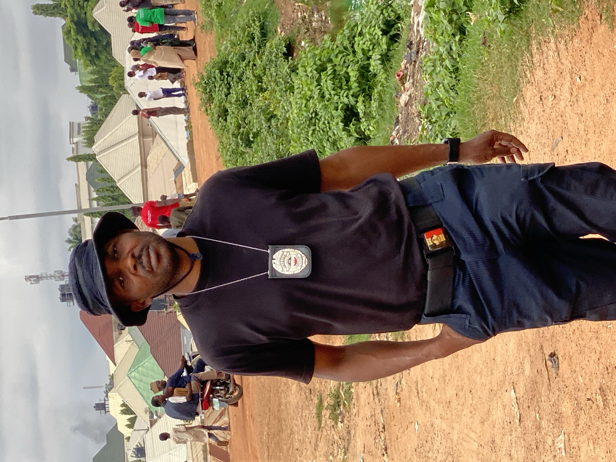 The image shows a man wearing a black bucket hat, a black t-shirt, and dark blue pants. He has a badge hanging from a chain around his neck and a belt with a buckle. His left hand is in his pocket. In the background, there are several buildings with pitched roofs, some appearing to be residential homes. There are also several people, some walking and others on a motorcycle. The ground is unpaved and dusty, with some patches of green vegetation and scattered debris. The sky is overcast. This scene is located in Kawo, Nigeria.