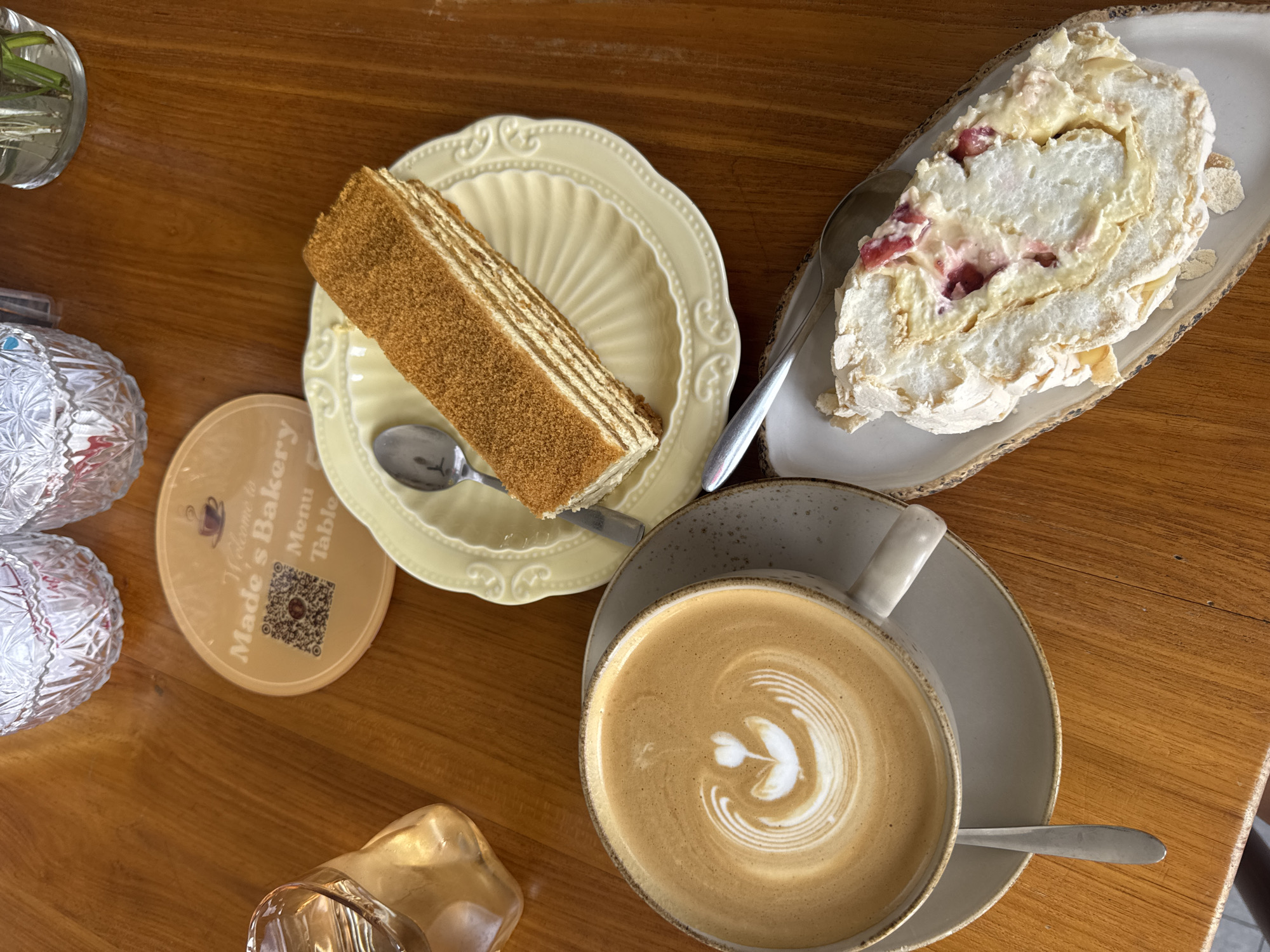 A overhead view shows a rustic wooden table at Made's Bakery in Kuta Selatan, Indonesia, set with a variety of treats and beverages. A cup of latte with intricate latte art rests on a saucer, accompanied by a spoon. To the right, a slice of honey cake is presented on a yellow, embossed plate, with a spoon resting on it. Below the cake, a roulade with meringue and berries is served on a pale ceramic dish. A small coaster with the bakery's logo and a QR code is visible on the table. A glass of water sits to the left, and decorative crystal glasses are in the background. The scene captures a moment of enjoying baked goods and coffee in a cozy cafe setting.