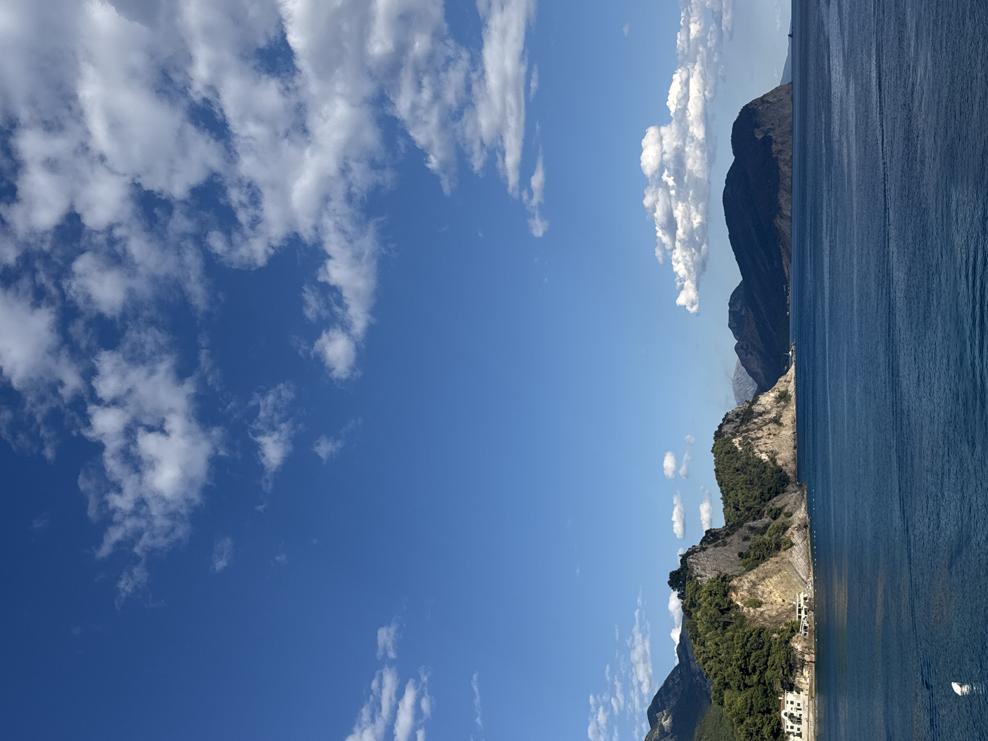 The image depicts a serene coastal scene in Petrovac centar, Montenegro. A vast expanse of deep blue sea dominates the lower half of the frame, with gentle ripples indicating calm waters. In the distance, a rugged coastline is characterized by steep, rocky cliffs covered in lush green vegetation. Several buildings, including what appears to be a white hotel or villa, are nestled along the shore. The background features a series of majestic mountains, some of which are shrouded in shadow, while others are bathed in sunlight. The sky above is a vibrant blue, dotted with fluffy white clouds, creating a picturesque and tranquil atmosphere.