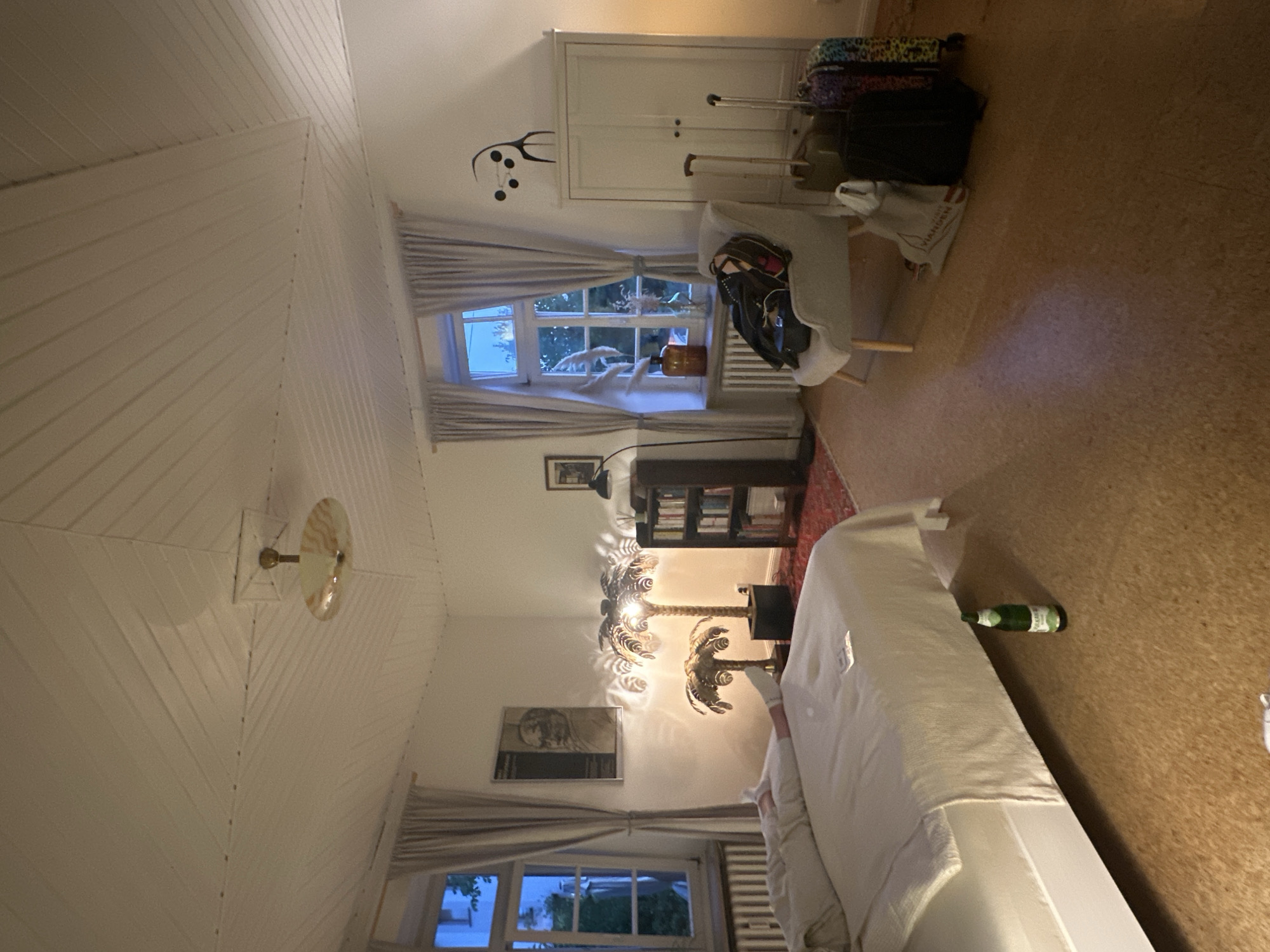 The image depicts a bedroom interior, possibly in a hotel or guesthouse, in Ammeldingen bei Neuerburg, Germany. A person is lying on a bed covered with white bedding, their legs extended and visible from the knees down, wearing white socks. The room features a sloped, wood-paneled ceiling with a circular light fixture. To the left, a window with light grey curtains is visible, and a radiator is beneath it. A framed picture hangs on the wall. In the center of the room, two palm-tree-shaped lamps illuminate the space. A dark bookshelf filled with books stands against the wall. To the right, a light grey armchair holds several bags, including a black jacket draped over the back. Adjacent to the armchair is a tall, white wardrobe with two doors. Several suitcases, including one with a colorful leopard print, are placed on the floor in front of the wardrobe. A green glass bottle lies on the cork floor near the bed. The overall lighting suggests it is evening or nighttime, with artificial light sources creating a warm ambiance.