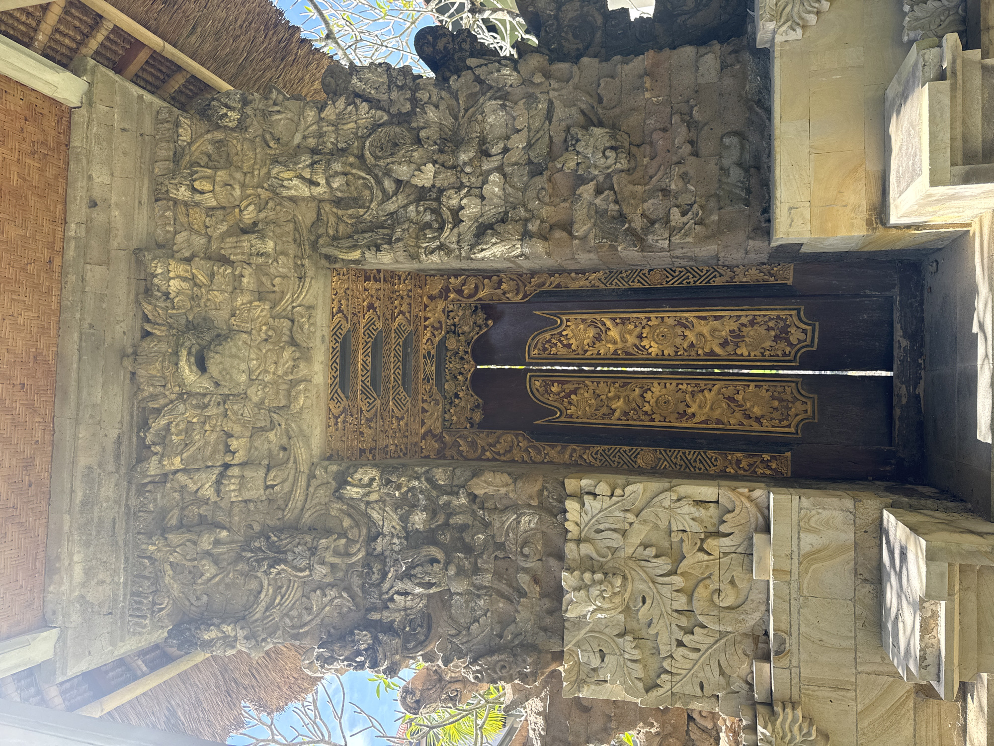 A dark, ornate wooden door is framed by intricate stone carvings. The carvings are highly detailed with floral and abstract motifs, suggesting a Balinese architectural style. The stone itself appears aged, with visible wear and variations in color. The door frame is set within a rough-hewn stone structure, partially covered by a thatched roof in the upper left corner. The environment suggests a location in Kuta Selatan, Indonesia, given the traditional architectural elements. No people are visible in the shot. The scene is bathed in natural light, with varying degrees of shadow highlighting the textured surfaces.