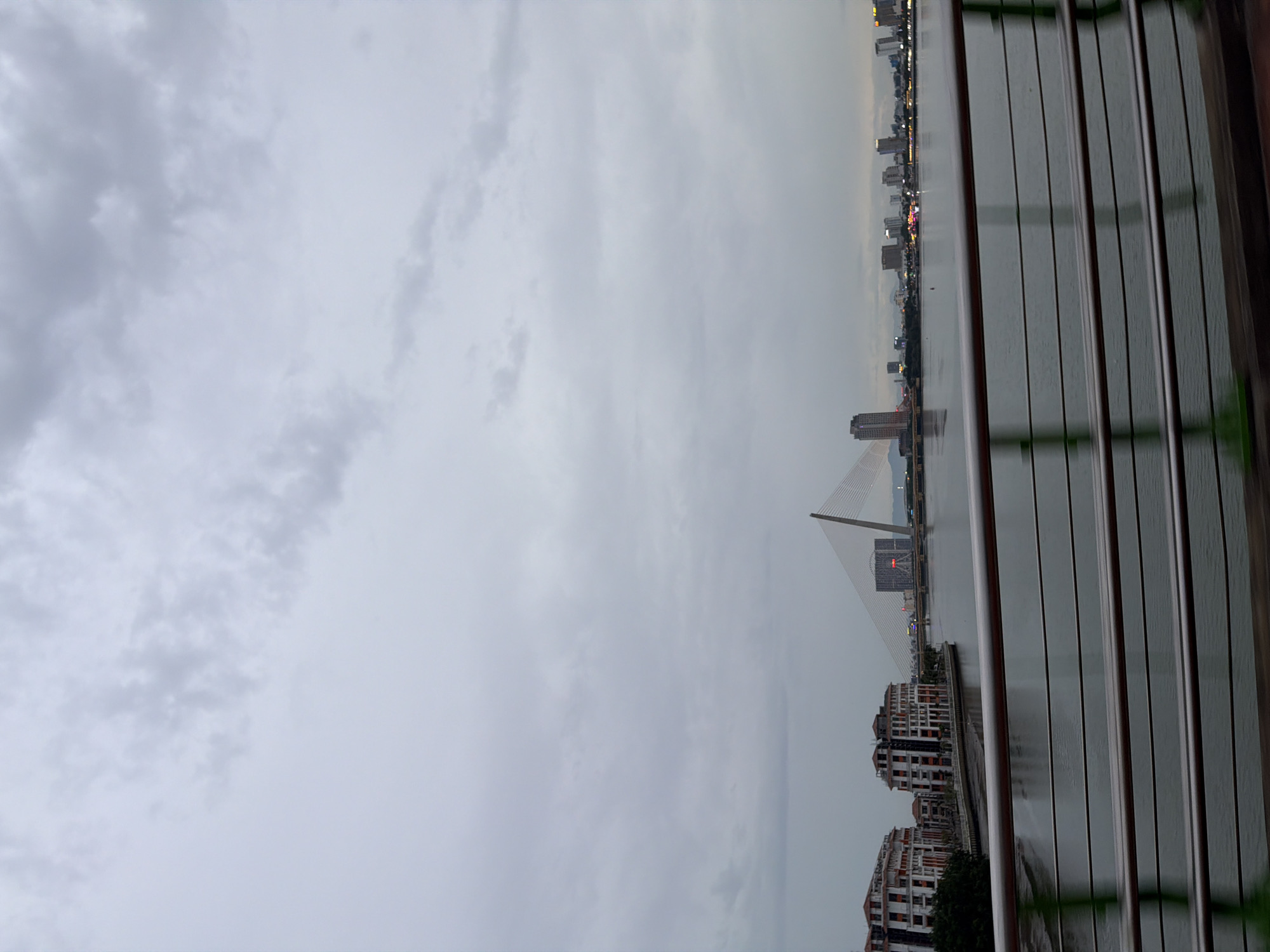 The image depicts a wide river with a modern cityscape on the far bank, viewed from what appears to be a bridge or elevated walkway. The sky is overcast and gray, with soft, diffused clouds. The water is calm, reflecting the muted light and the structures along the riverbank.
On the left side of the riverbank, there are several multi-story buildings with white facades and red-tiled roofs, some appearing to be residential or commercial complexes. A cluster of trees is visible behind these buildings.
Dominating the center of the far bank is a striking cable-stayed bridge with a single, tall, angled pylon. In the distance, behind the bridge, a Ferris wheel is illuminated against the twilight sky. Further along the cityscape, various modern buildings and skyscrapers rise, some with lights visible. The overall impression is one of urban development and modern infrastructure.
This scene is identifiable as the Han River in Đà Nẵng, Vietnam, specifically showing the Dragon Bridge (the cable-stayed bridge) and the surrounding city skyline. The perspective suggests the photo was taken from a vantage point looking east, potentially from the Trần Thị Lý Bridge or a similar elevated structure. No people are clearly visible in the frame, and the focus is on the urban landscape and the river.