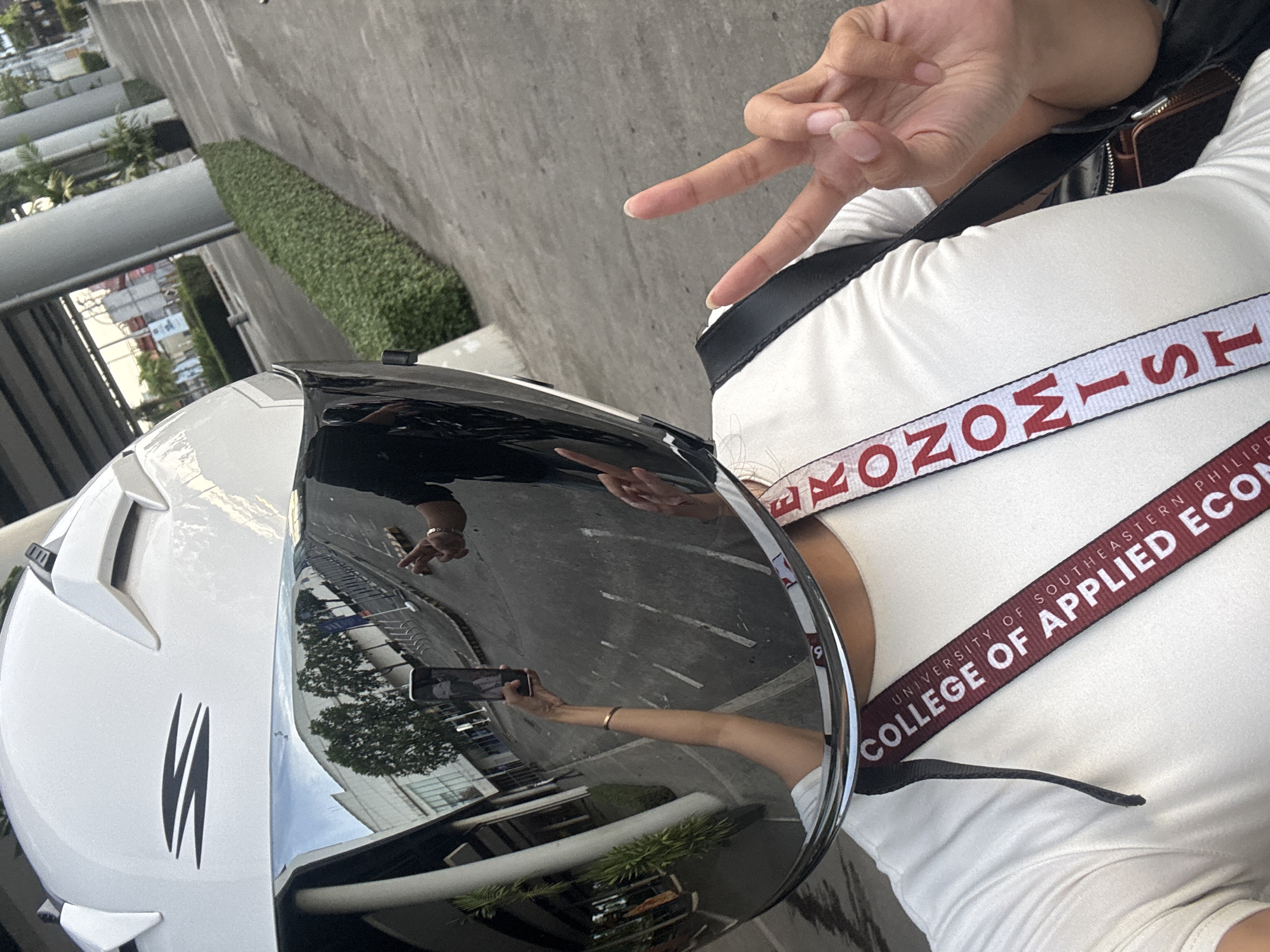 The image presents a first-person perspective, likely a selfie. The subject is wearing a white, long-sleeved shirt, with two lanyards/straps across their chest. One lanyard has the text "UNIVERSITY OF SOUTHEASTERN PHILIPPINES COLLEGE OF APPLIED ECON", and the other reads "ECONOMIST." The subject is also wearing a motorcycle helmet with a highly reflective visor. The visor reflects a scene of a city street, with buildings and foliage visible. Another hand is reflected in the visor, holding a smartphone taking the photo. The background outside the visor includes a concrete wall, a hedge, and part of a building. It may be taken in Davao City, Philippines, based on the lanyard text. The subject is making a "peace" sign with their fingers.