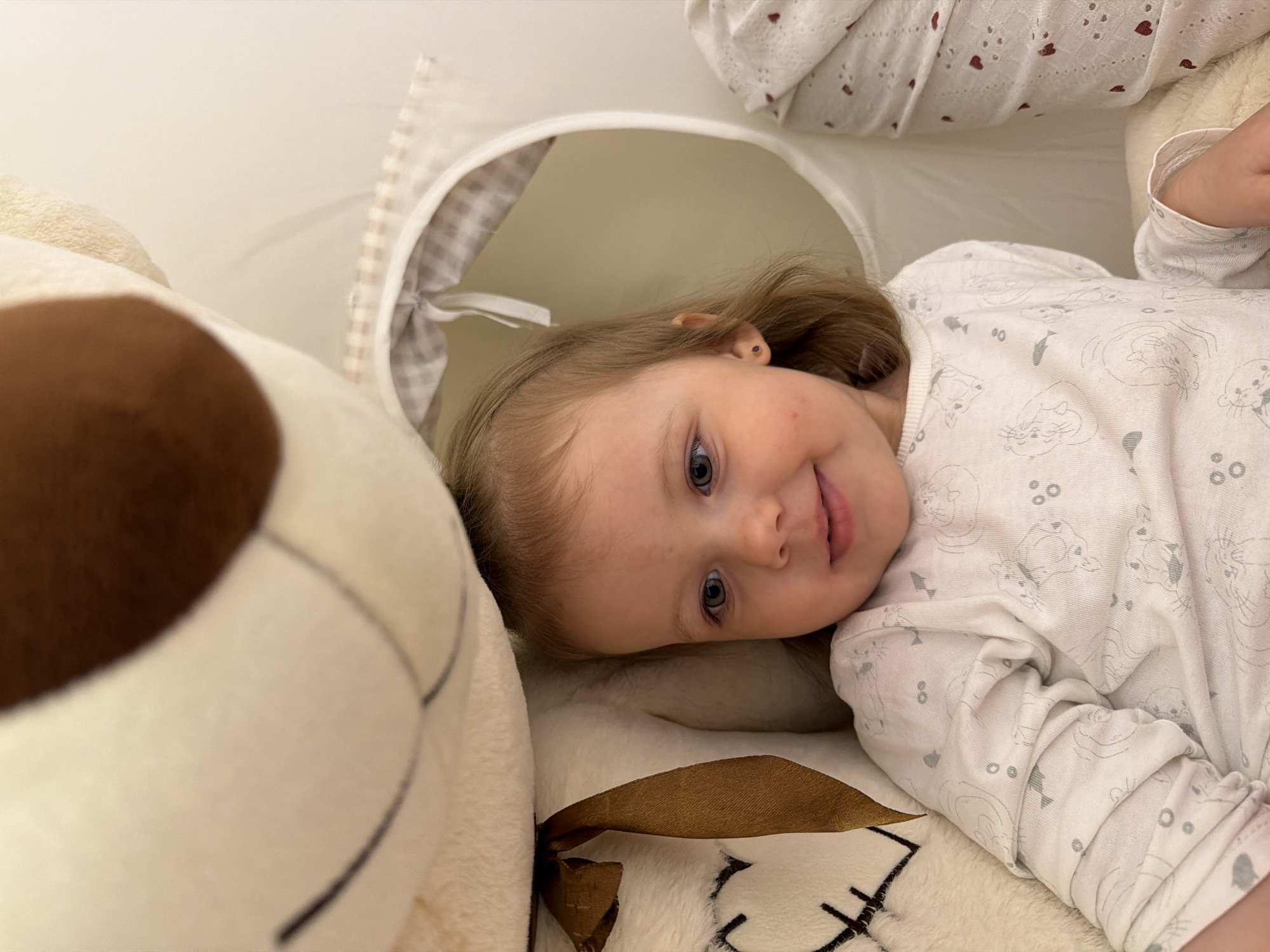 A young child is lying on a soft, white surface, possibly a bed or play area. The child is wearing white pajamas with grey animal prints. A large, plush teddy bear with a brown nose is positioned to the left of the child. Behind the child's head is a white, circular structure with a checkered grey lining, possibly a toy tunnel or play structure. The background is primarily white, suggesting an indoor setting. There is no information available to confirm if this image was taken in the city of Reshetnikovo, Russia.