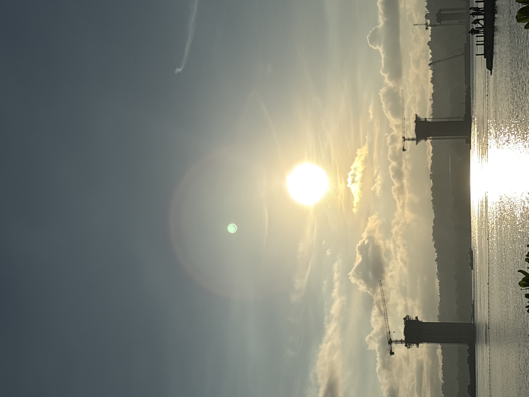 The image captures a panoramic view of a construction site on the coast of Davao City, Philippines, likely near the Samal Ferry Terminal or the ongoing bridge project. The sun is prominently positioned in the sky, casting a bright, diffused light that creates a halo effect and glints on the water's surface. The sky is a gradient of soft blues and grays, with wispy clouds scattered across it.
In the foreground, the water is a calm expanse, reflecting the bright sunlight. Along the water's edge, there is a hint of greenery, possibly plants. Further out, a boat is visible, moving across the water.
The middle ground of the image is dominated by the construction of what appears to be a bridge or a large infrastructure project. Two towering concrete pillars, topped with cranes, stand silhouetted against the sky. These structures are under active construction, with the cranes indicating ongoing work. Behind these pillars, a distant, tree-covered shoreline forms the backdrop of the scene.
On the right side of the image, a small pier extends into the water. On this pier, a group of four to five individuals can be seen, appearing to be observing the construction or the sunset. Their figures are small in the frame, suggesting they are distant. The overall impression is one of industrial development set against a natural, serene coastal environment.