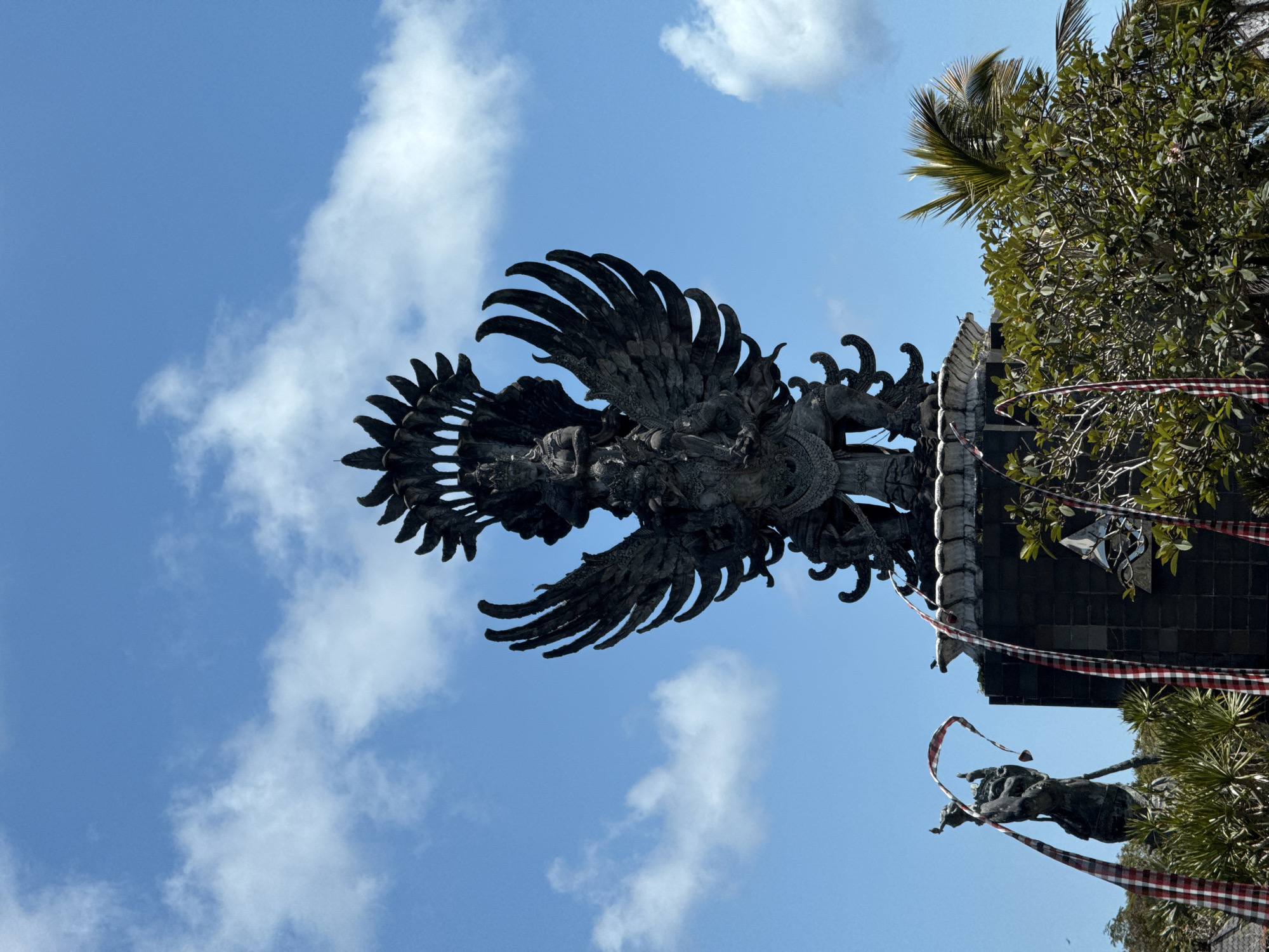 The image, taken in Kuta Selatan, Indonesia, captures a grand, dark-colored stone or metal statue of a mythical winged figure, likely a Garuda, soaring against a clear, bright blue sky dotted with scattered white clouds. The statue is intricately carved, featuring expansive, feathered wings and a detailed humanoid torso, conveying a sense of majesty and power.
To the right of the prominent winged statue, a traditional Balinese architectural structure, possibly a temple gate or part of a larger complex, is visible. It features dark, stone-like elements and ornate decorative patterns at its upper edge. Lush green foliage, including palm fronds and dense tree leaves, frames the right side of the scene, partially obscuring the structure and adding a natural element to the man-made art.
Below the main structure and foliage, a smaller, dark statue or carving is partially visible. Draped prominently across the scene, hanging from the architecture and near the smaller statue, are long, checkered red and white ribbons, characteristic of Balinese "poleng" fabric, often used in religious ceremonies or as protective symbols. The bright daylight suggests it is daytime with good weather. There is no visible text.