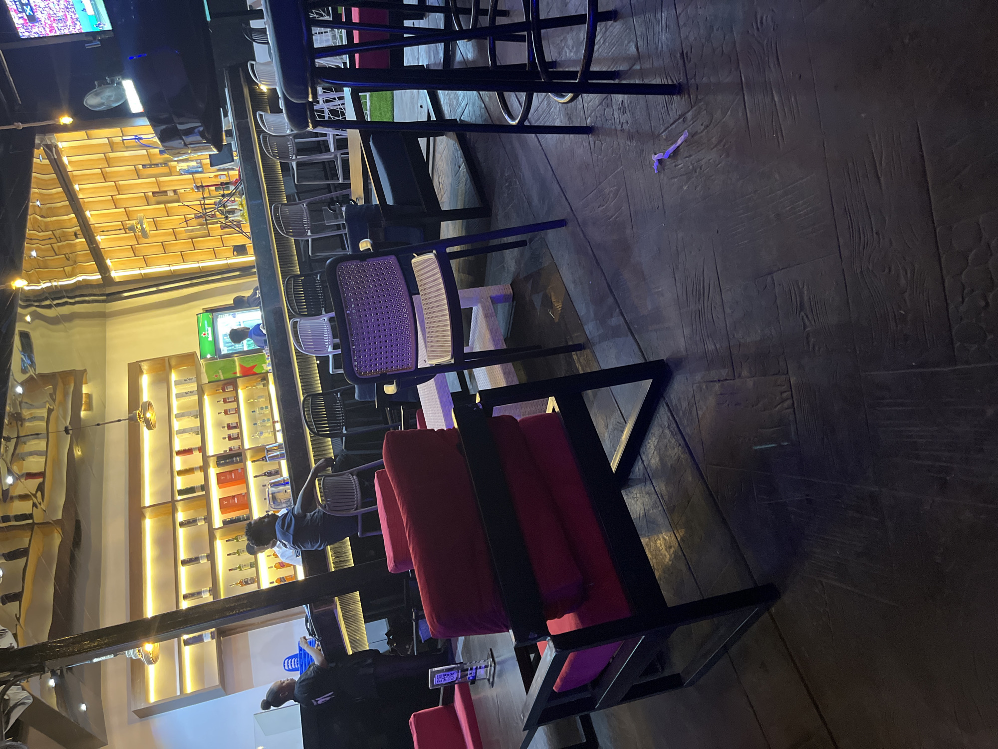 The image depicts the interior of a bar or lounge in Abuja, Nigeria. In the foreground, there are several chairs and tables. A prominent dark-framed chair with red cushions is in the lower left, facing towards the right. To its right, a black chair with a woven seat and back is positioned. Beyond these, more seating and tables are visible, including a long black sofa with dark cushions and a white woven chair.
In the middle ground, a bar area is illuminated by warm yellow lighting. Behind the bar, shelves are stocked with various bottles of alcohol. Two individuals are visible behind the bar. One, wearing a dark top, appears to be a server or bartender, interacting with a customer. Another person, wearing a blue shirt, is also behind the bar. Several bar stools are arranged along the counter. A green refrigerator with a beer logo is situated on the left side of the bar.
The background features a stylish, modern design with wooden paneling and reflective surfaces on the ceiling, suggesting a contemporary establishment. The flooring in the foreground appears to be dark wood planks with a textured finish. The overall atmosphere suggests a relaxed, social environment.