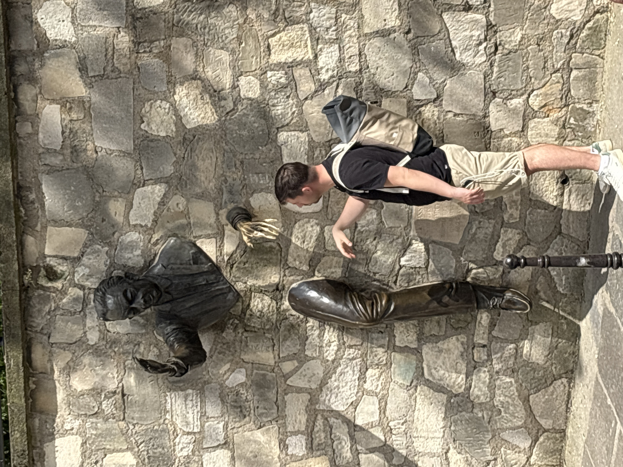 The image captures a bright, sunny daytime scene in Paris, France, showing a man interacting with a unique bronze sculpture embedded in a stone wall. The man, dressed in a black t-shirt, khaki shorts, and white sneakers, with a beige and black backpack, is leaning towards the wall and extending his right hand.
The sculpture, known as "Le Passe-Muraille" (The Walker Through Walls) in Montmartre, depicts a man appearing to emerge from the rough, irregular stone wall. The bronze figure's upper body, including his head and arms, is visible, portraying a pensive or thoughtful expression. His left hand is open and outstretched, while his right hand appears to hold a small object. Additionally, a separate bronze arm and hand are seen slightly above the main figure's arm, and a bronze leg from the knee down, complete with a shoe, protrudes from the wall below the main torso.
Strong shadows cast by the figures and the man indicate bright sunlight, suggesting it is likely mid-day or afternoon. The wall is constructed from light-colored, irregularly shaped stone blocks. A dark metal railing or post is partially visible on the far right. The overall scene depicts a person engaging with a public art installation, possibly posing for a photograph. No visible text is present in the image.