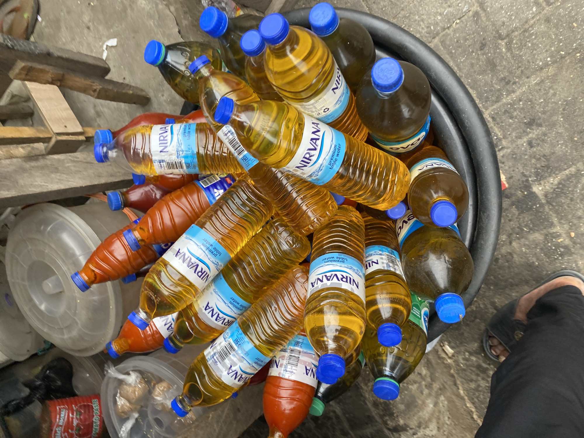 The image depicts an assortment of plastic bottles, mostly filled with liquids of varying hues ranging from yellow to orange, clustered in a large black bucket. The bottles bear labels predominantly featuring the word "NIRVANA" and "Premium table water." Some bottles also bear the brand name "EVA." A few bottles contain a reddish-orange liquid.
The immediate environment surrounding the bucket includes a haphazard arrangement of what appears to be wooden planks or parts of furniture, along with various plastic containers of different sizes. There are hints of food packaging and food items. The ground surface is characterized by a combination of concrete and what looks like a gray, uneven street or floor.
The presence of a leg clad in black pants and a sandal-clad foot places an individual nearby, although their actions or interactions with the objects are not discernible. The scene, based on the branding of the water bottles and the general urban setting, suggests a location in Lekki, Nigeria. The composition of the scene gives an impression of a recycling or storage area for used beverage containers.
